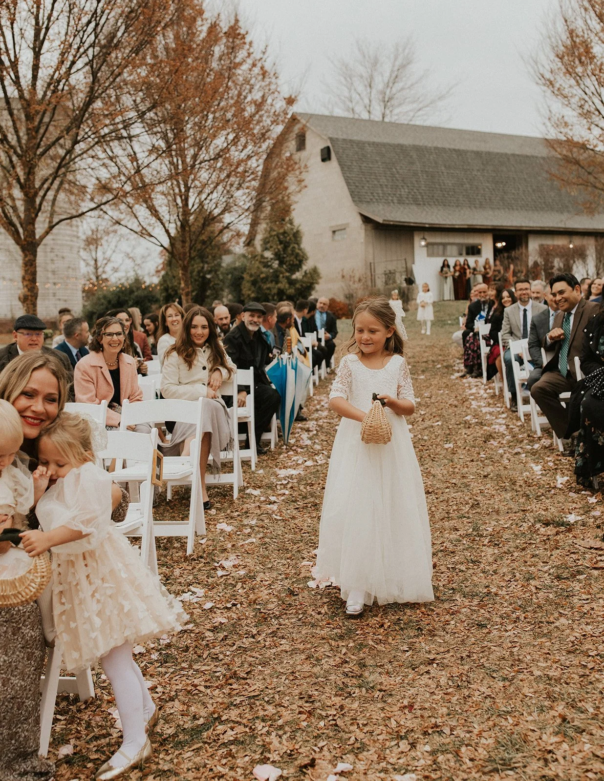 Children and adults at an outdoor wedding ceremony with leaf-covered ground, trees with autumn leaves, and a rustic barn in the background.