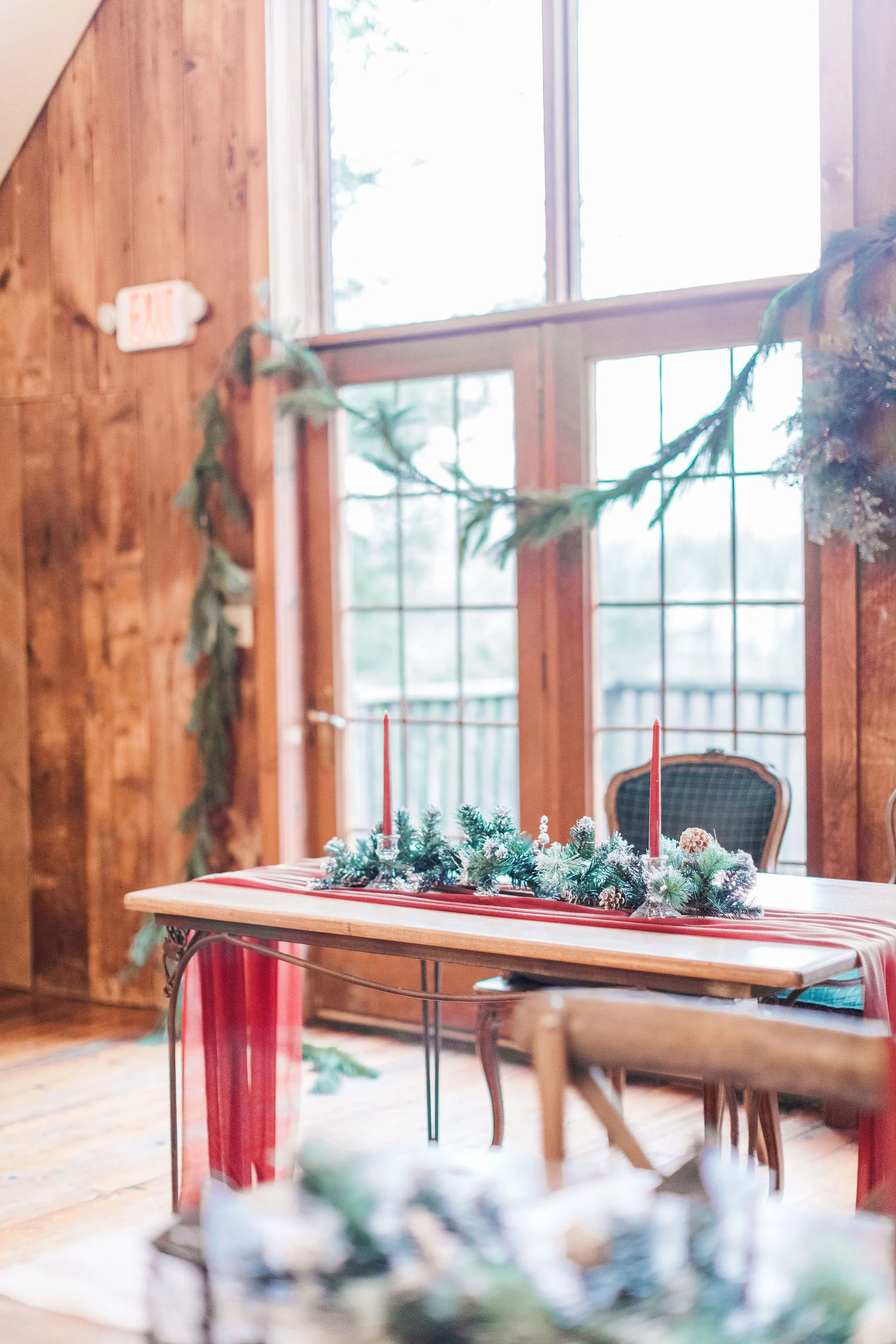 A wooden table with a Christmas centerpiece featuring greenery, pinecones, and red candles. The background shows wooden walls, large windows, and chairs, indicating a cozy indoor setting.