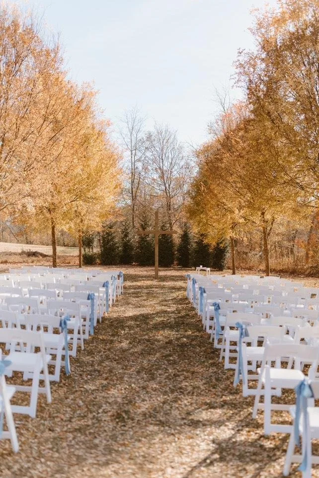 Outdoor wedding setup with white chairs decorated with blue ribbons arranged in rows along a grassy aisle, flanked by trees with autumn-colored leaves, and a wooden cross at the end of the aisle.