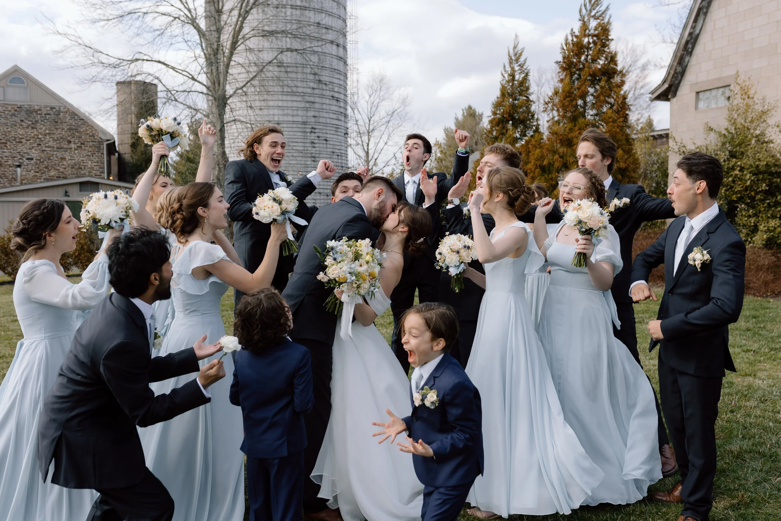A group of people in wedding attire celebrating outdoors, including a bride and groom kissing, surrounded by friends holding flowers and cheering.