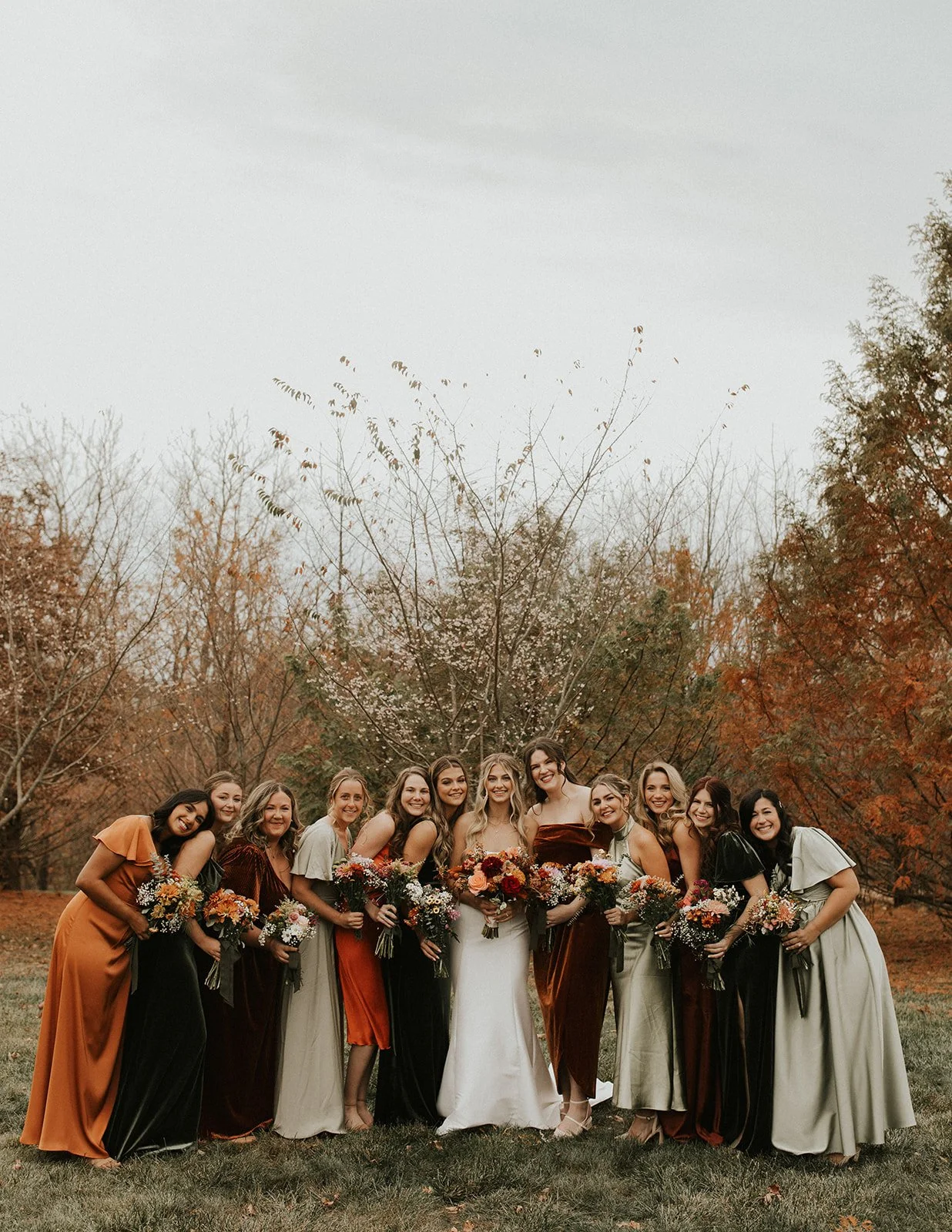 Group of women in formal dresses standing outdoors during fall, holding bouquets of flowers, smiling.