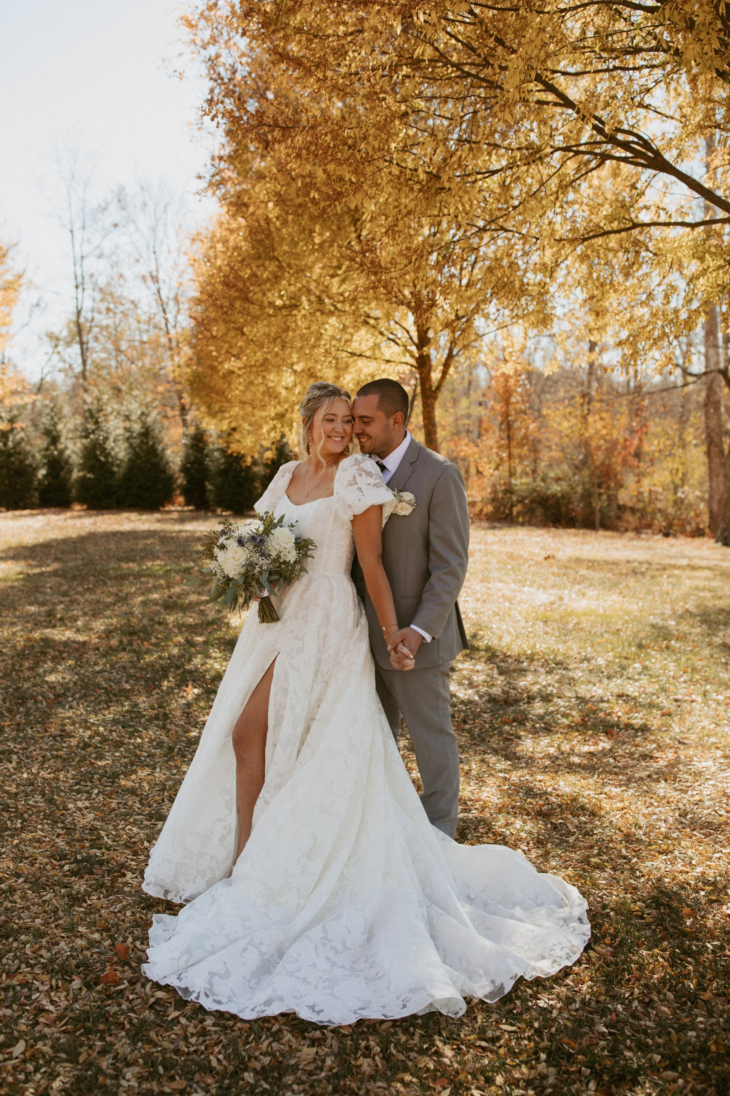 A bride and groom stand in an autumnal park, smiling and holding hands, surrounded by gold and orange fall foliage, with the bride holding a bouquet of white and purple flowers.