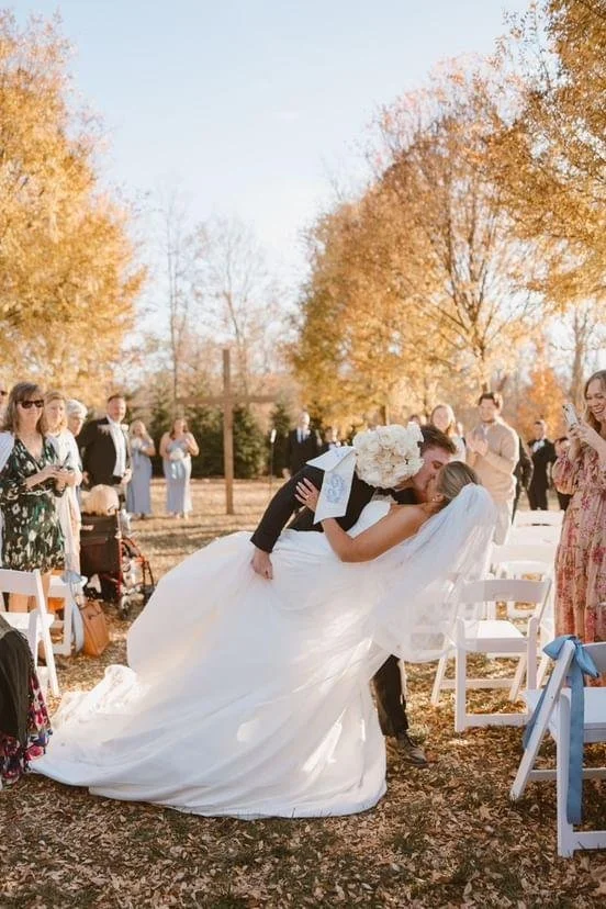 A bride and groom share a kiss at their outdoor wedding ceremony surrounded by guests and autumn trees.
