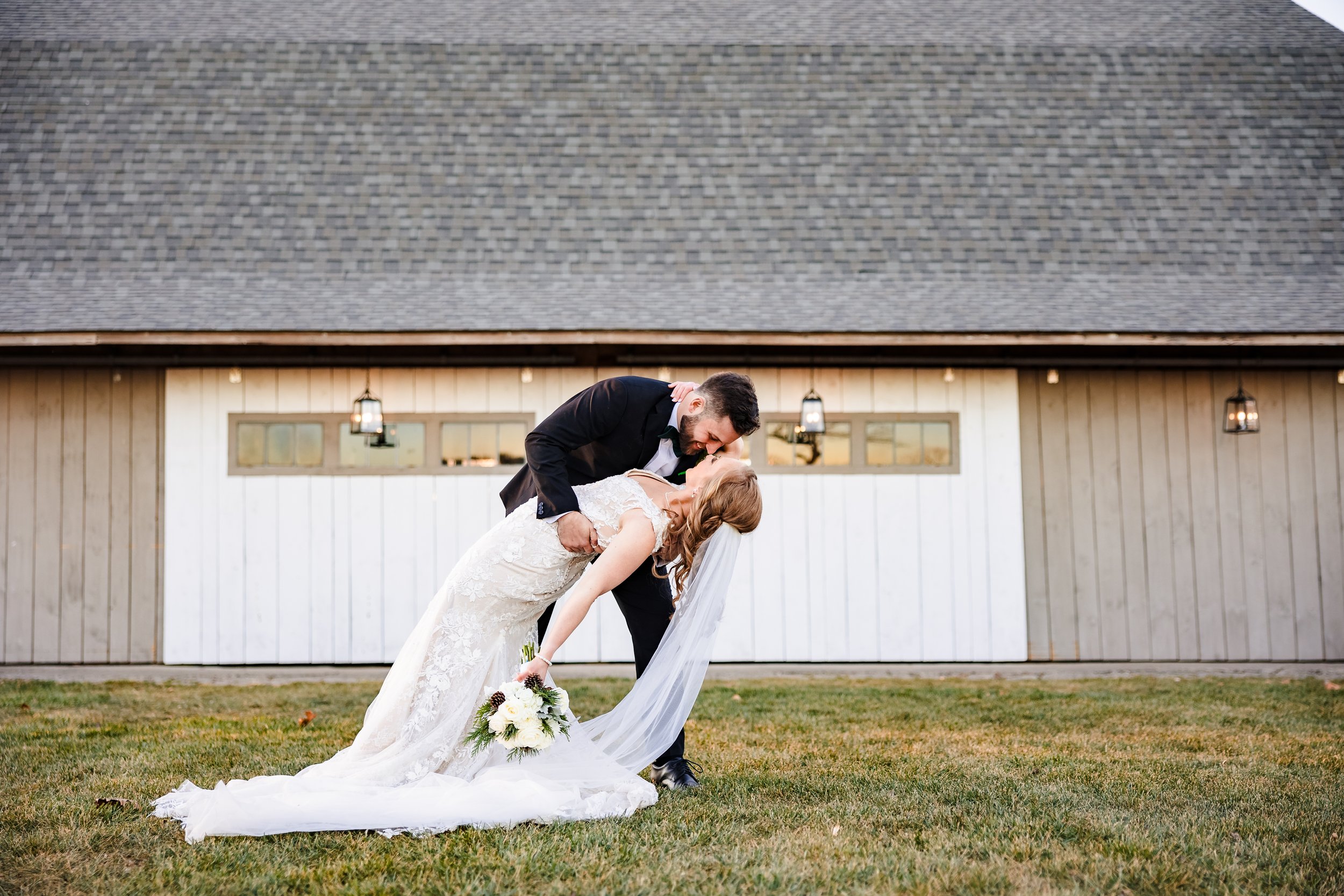 A groom and bride are dancing outdoors in front of a building with hanging lanterns, with the groom dipping the bride back and kissing her, holding her waist and her hand.
