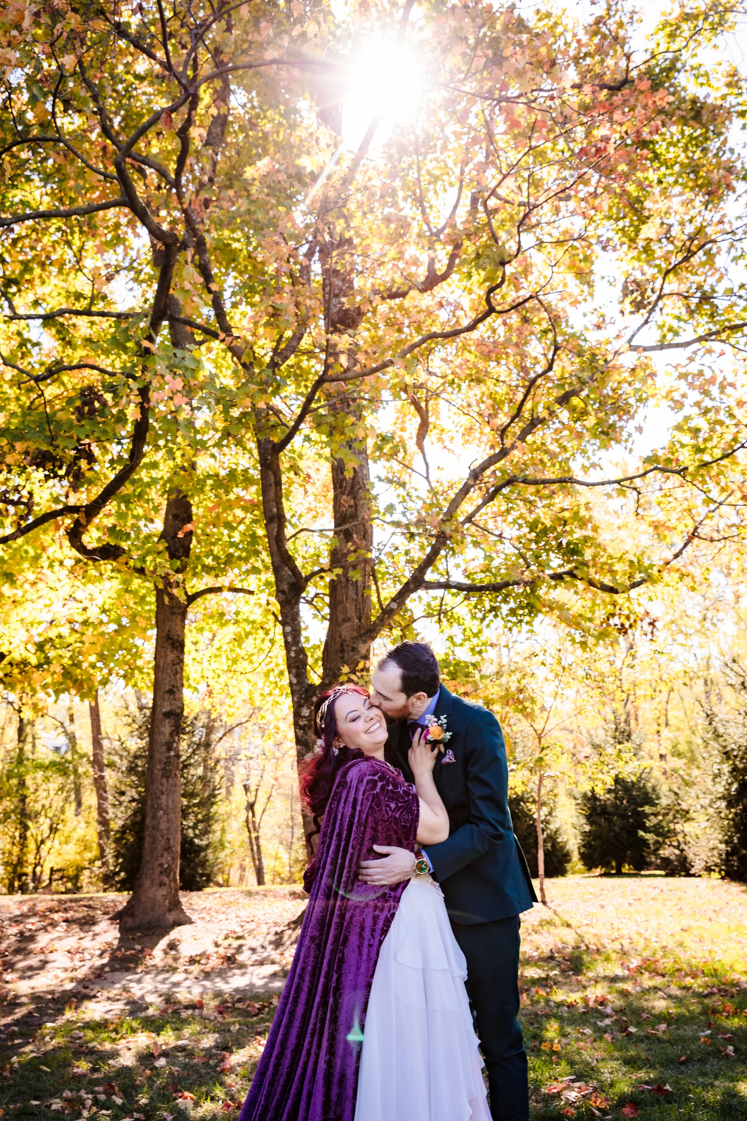 A couple, a woman with purple hair and a man in a tuxedo, embracing and sharing a kiss in a park during autumn. The sunlight filters through the colorful leaves on trees, creating a warm and romantic atmosphere.