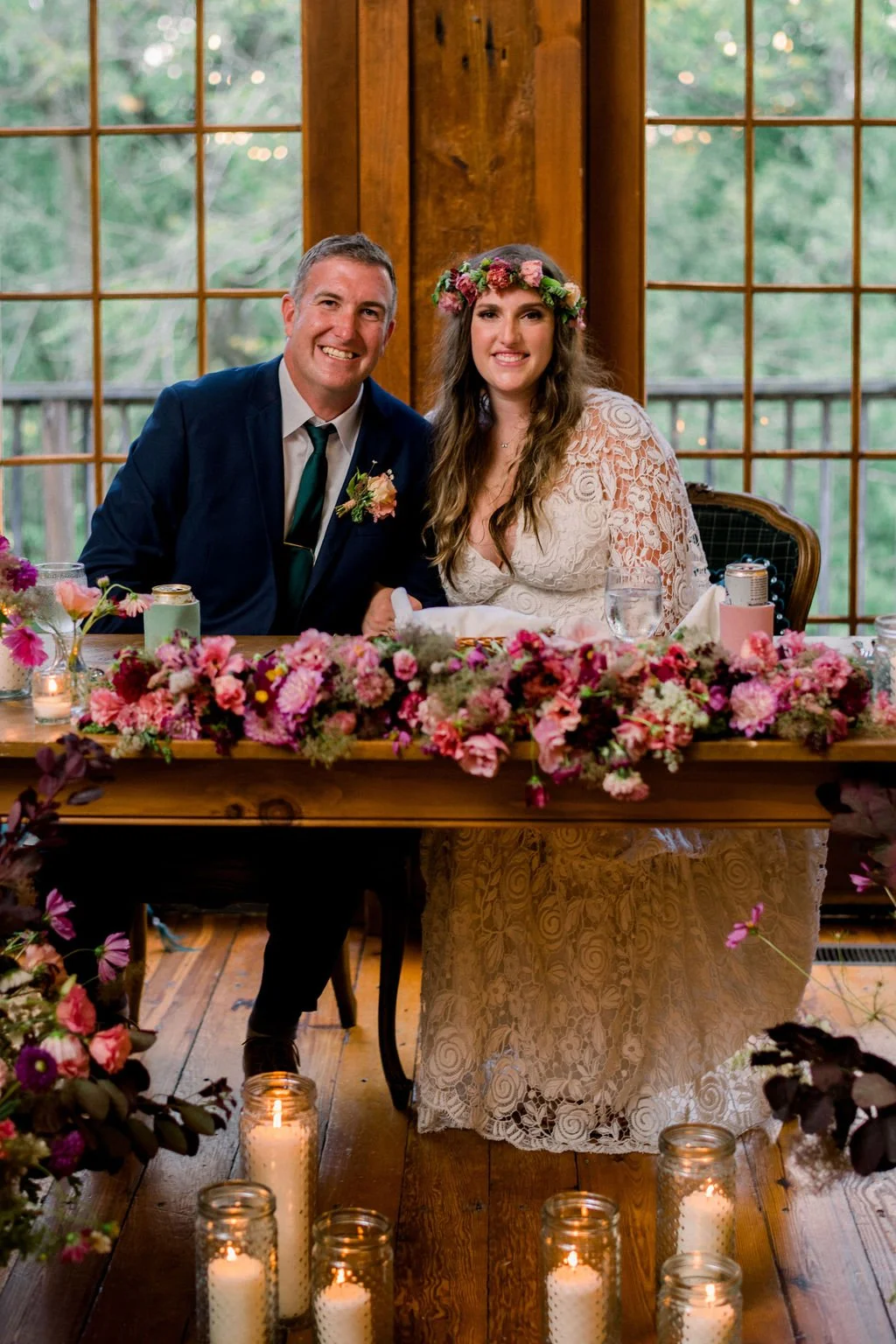 A bride and groom sitting at a decorated table during a wedding reception with wooden walls and large windows showing greenery outside. The table is adorned with pink and purple flowers and lit candles in glass jars.