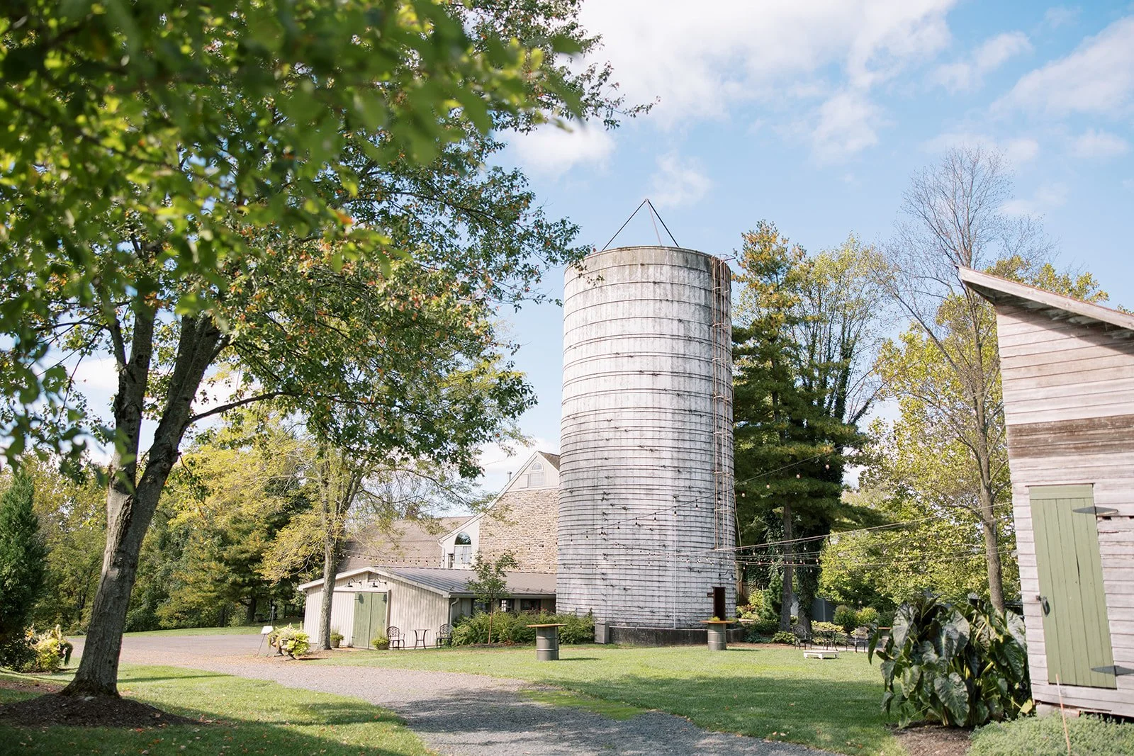 A white cylindrical silo surrounded by trees and small buildings under a partly cloudy sky.