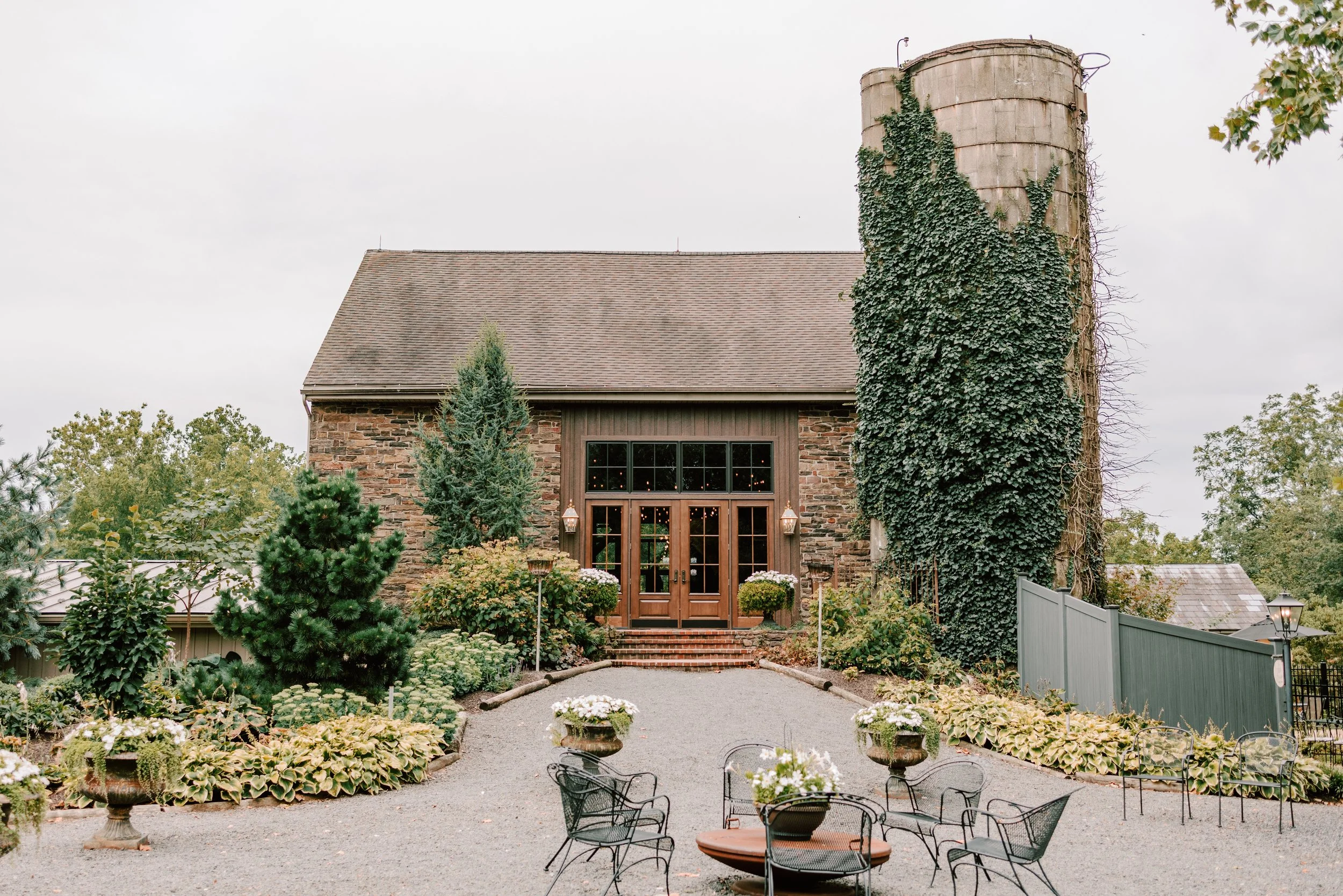 A rustic stone building with large glass doors and string lights inside, surrounded by a lush garden with various plants, flowers, and trees, and outdoor seating with black metal chairs in the foreground.