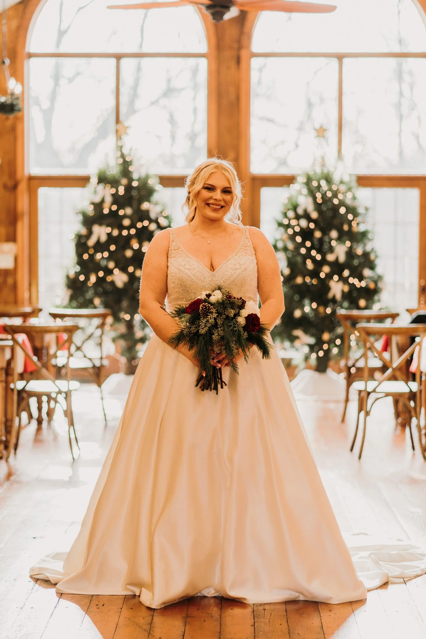A smiling bride in an ivory wedding gown holding a bouquet of red and white roses with pine branches, standing in a decorated venue with Christmas trees and large windows in the background.