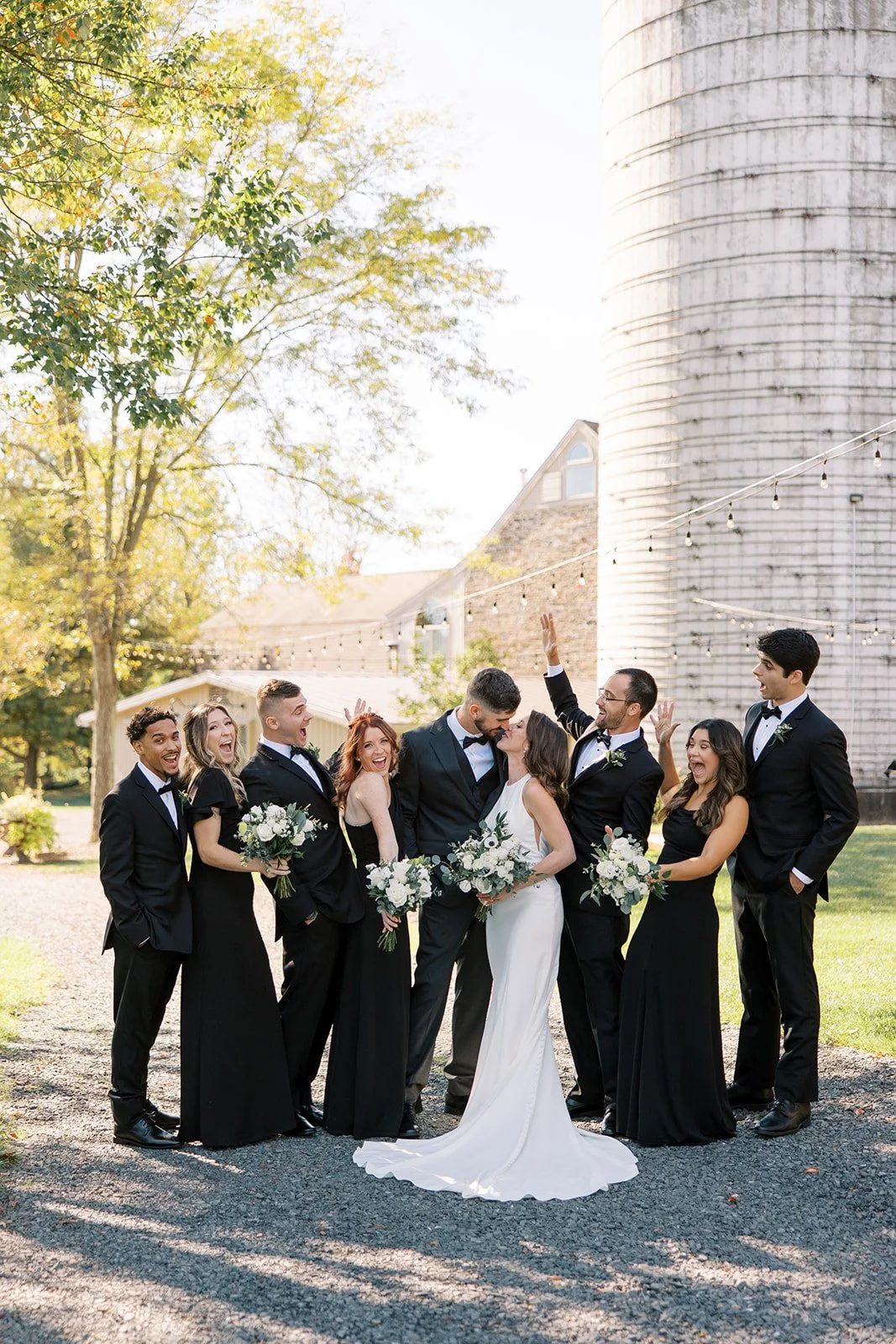 A wedding party with couples and friends standing outdoors on sunny day, celebrating and smiling, with a barn and trees in the background.