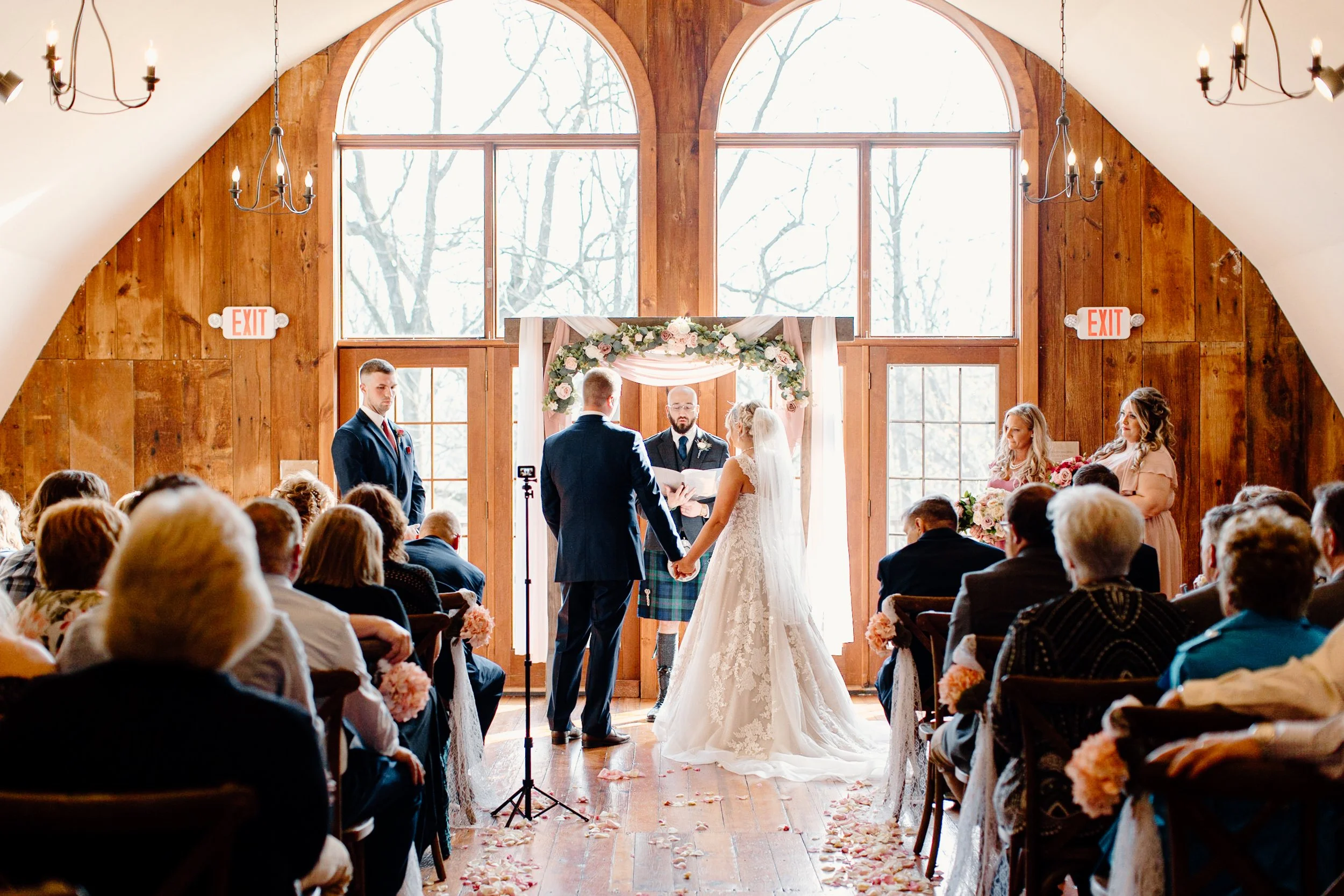A wedding ceremony taking place in a wooden chapel with large windows, where a bride and groom are holding hands under an arch with flowers. Guests are seated in rows, and a officiant stands behind them with a person recording the event.