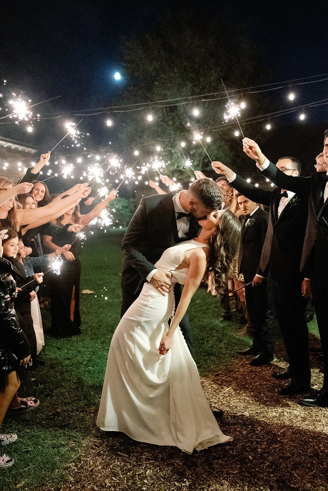 A bride and groom share a kiss at night during their wedding celebration, surrounded by guests holding sparklers, under a partly cloudy moonlit sky.
