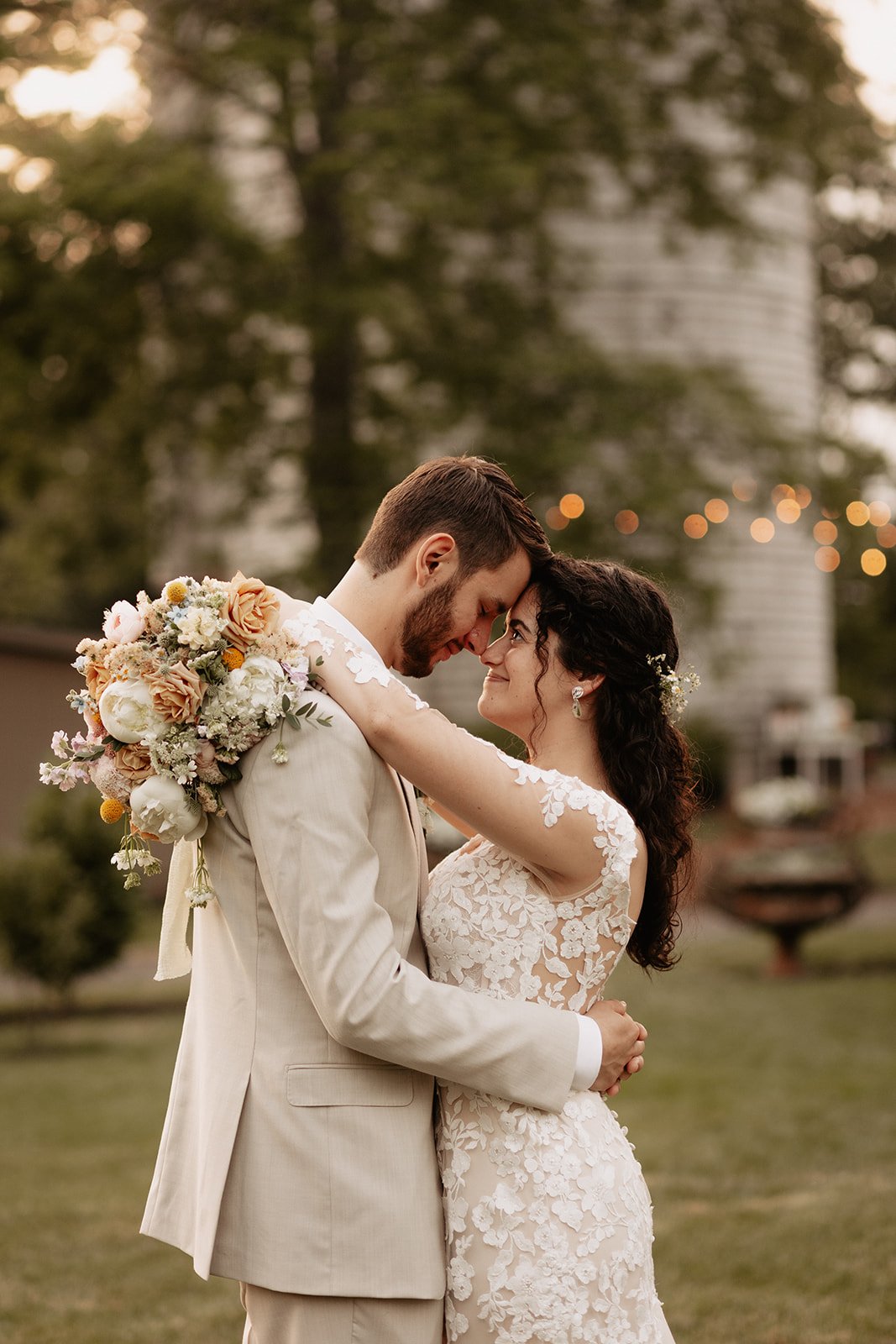 A newlywed couple embracing outdoors during sunset, with the bride in a lace wedding dress and the groom in a beige suit, holding a large bouquet of flowers.