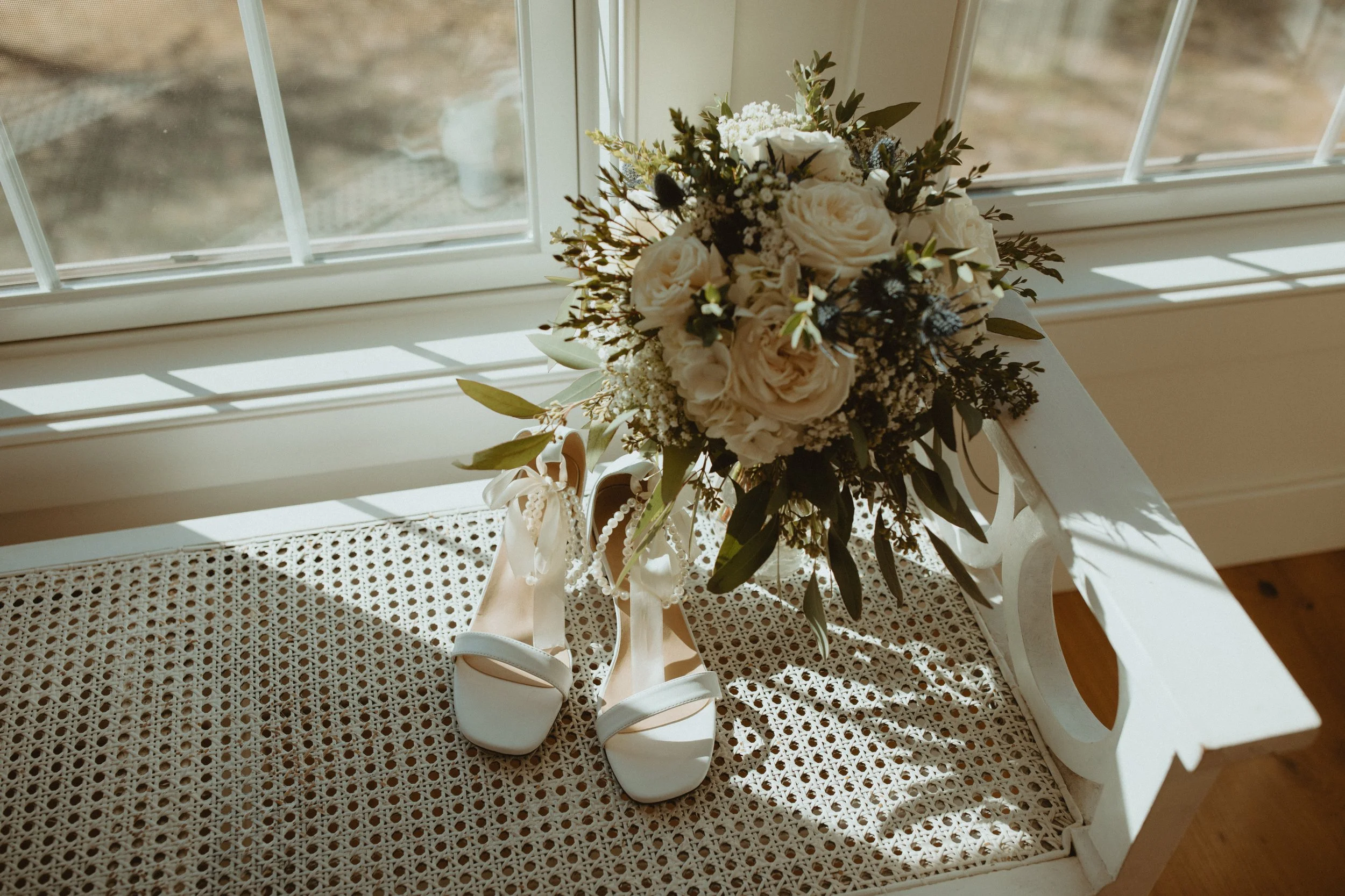 A bouquet of white and cream-colored flowers sits on a white wooden bench near a window with sunlight casting shadows.