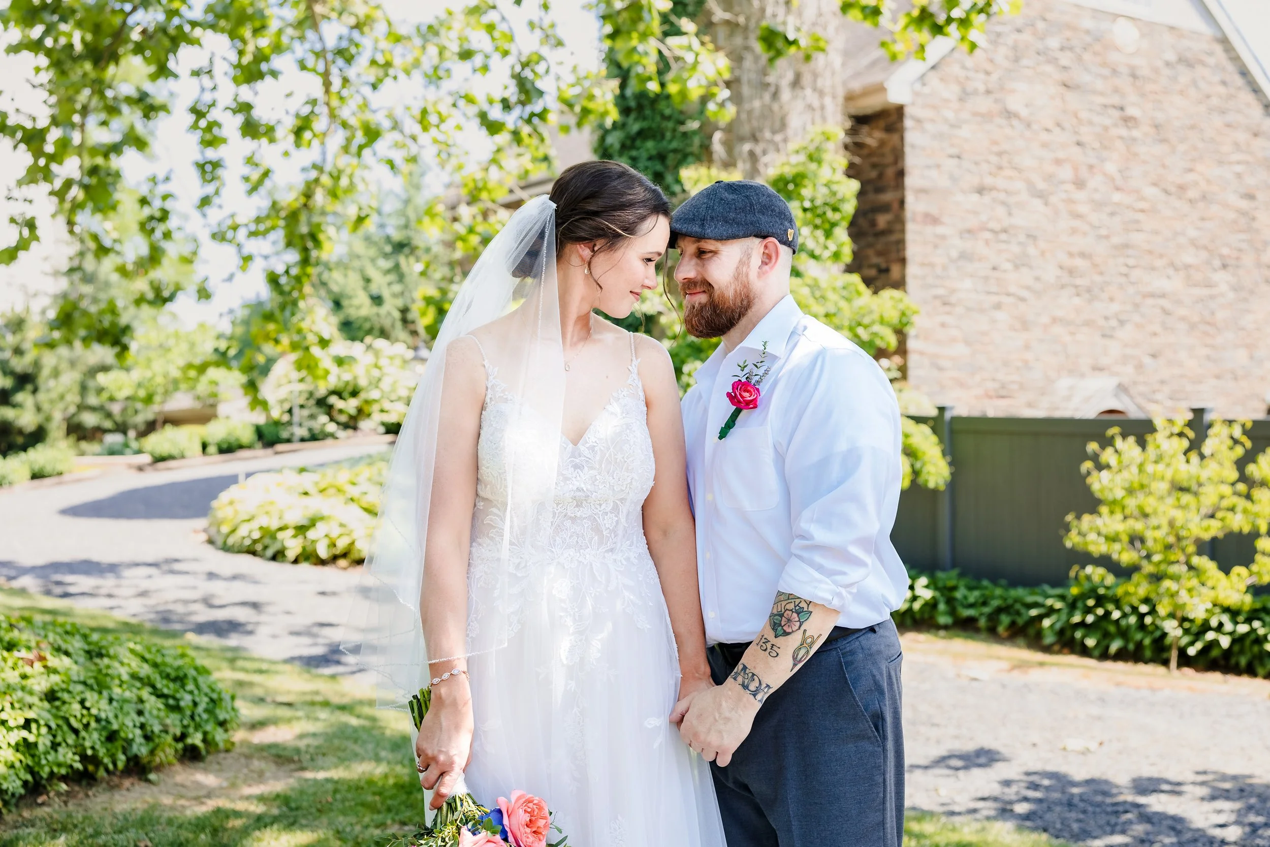 A bride and groom stand close together outdoors, touching foreheads and holding hands. The bride wears a white lace wedding dress and veil, holding a bouquet of flowers. The groom has a beard, tattoos on his arms, and wears a white shirt with a pink 