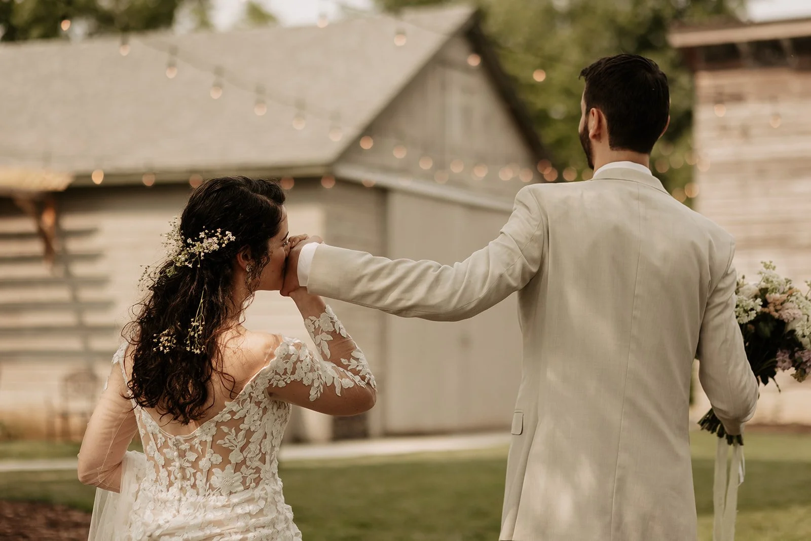 A groom in a light-colored suit is holding a bride's hand while wiping away a tear during an outdoor wedding ceremony. The bride is wearing a lace wedding dress with floral details and has flowers in her hair. The scene has soft lighting and a blurre