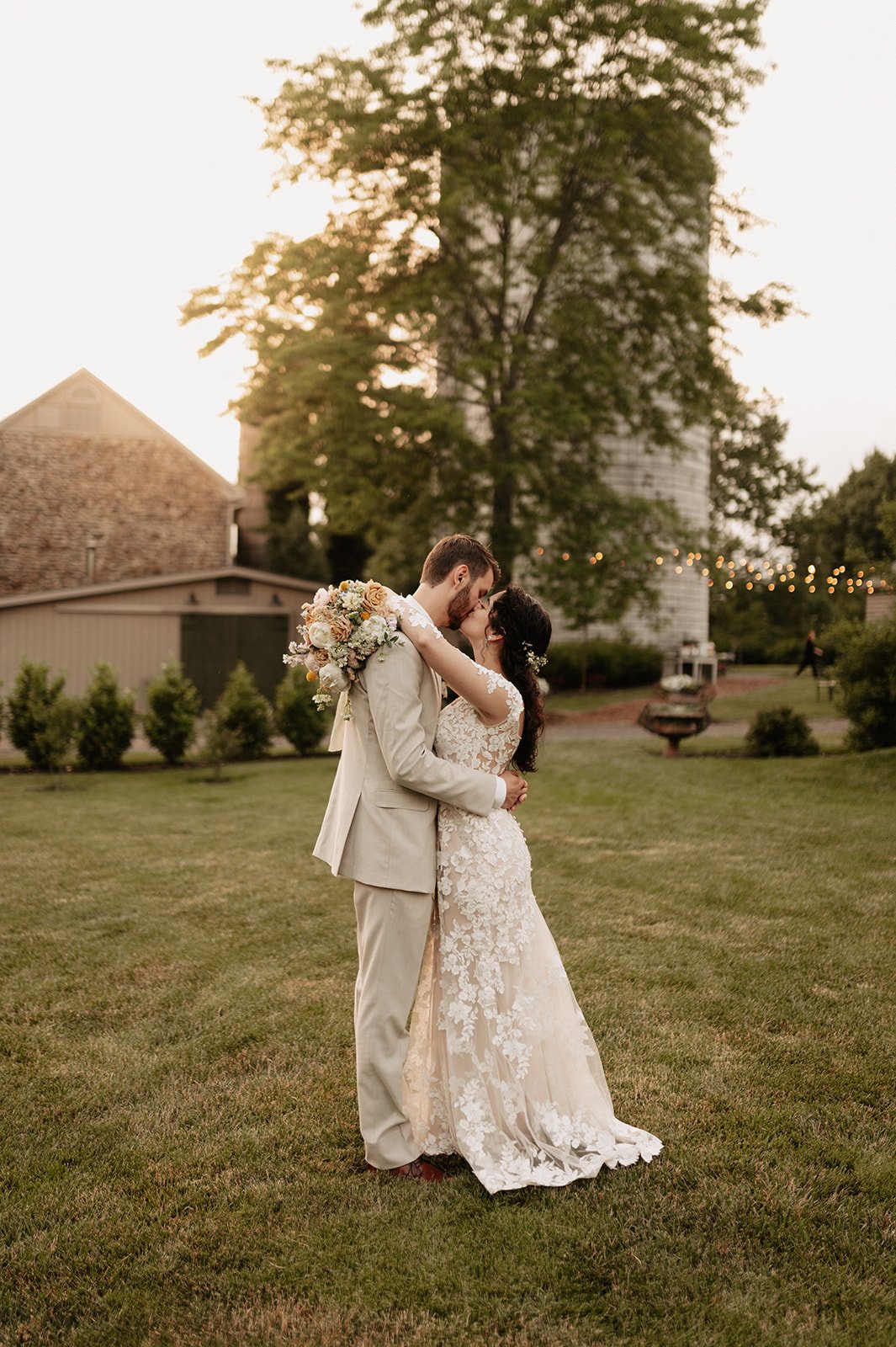 A newlywed couple sharing a kiss outdoors during sunset. The bride is in a lace wedding gown holding a bouquet, and the groom is in a light-colored suit.