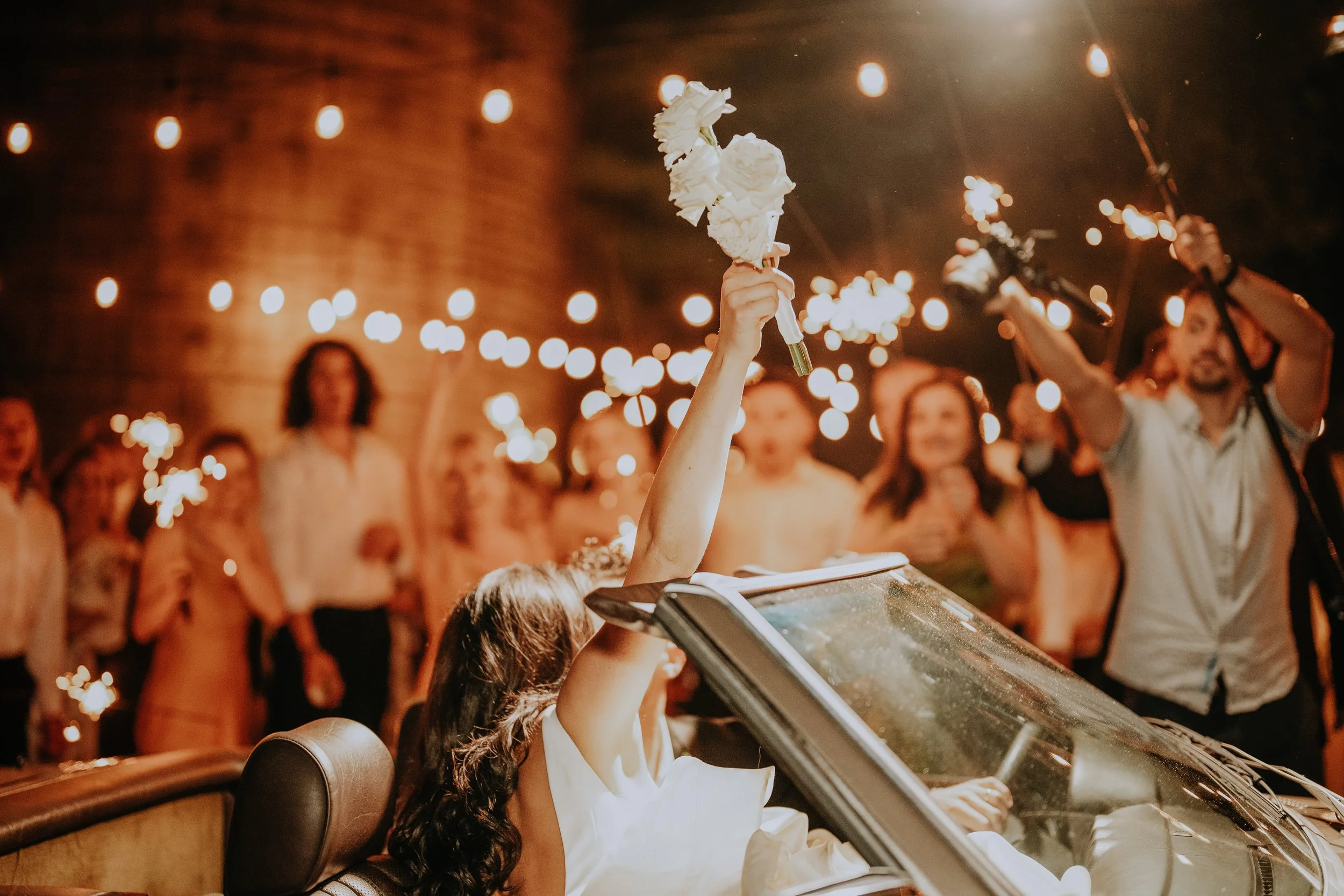 Person holding a white bouquet of flowers at a celebration with sparklers and a crowd in the background during evening time.