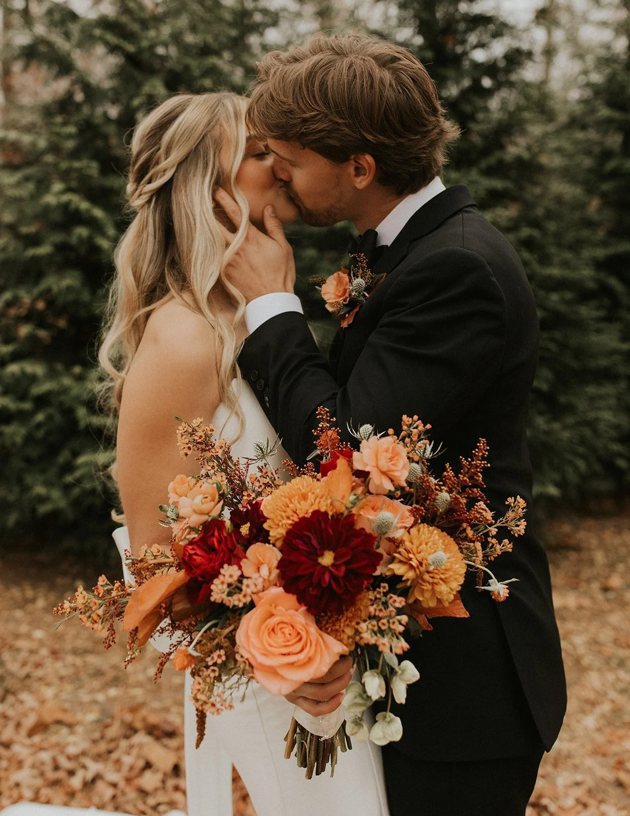 A bride and groom are kissing outdoors, with the groom holding a large orange, red, and peach bouquet of flowers.