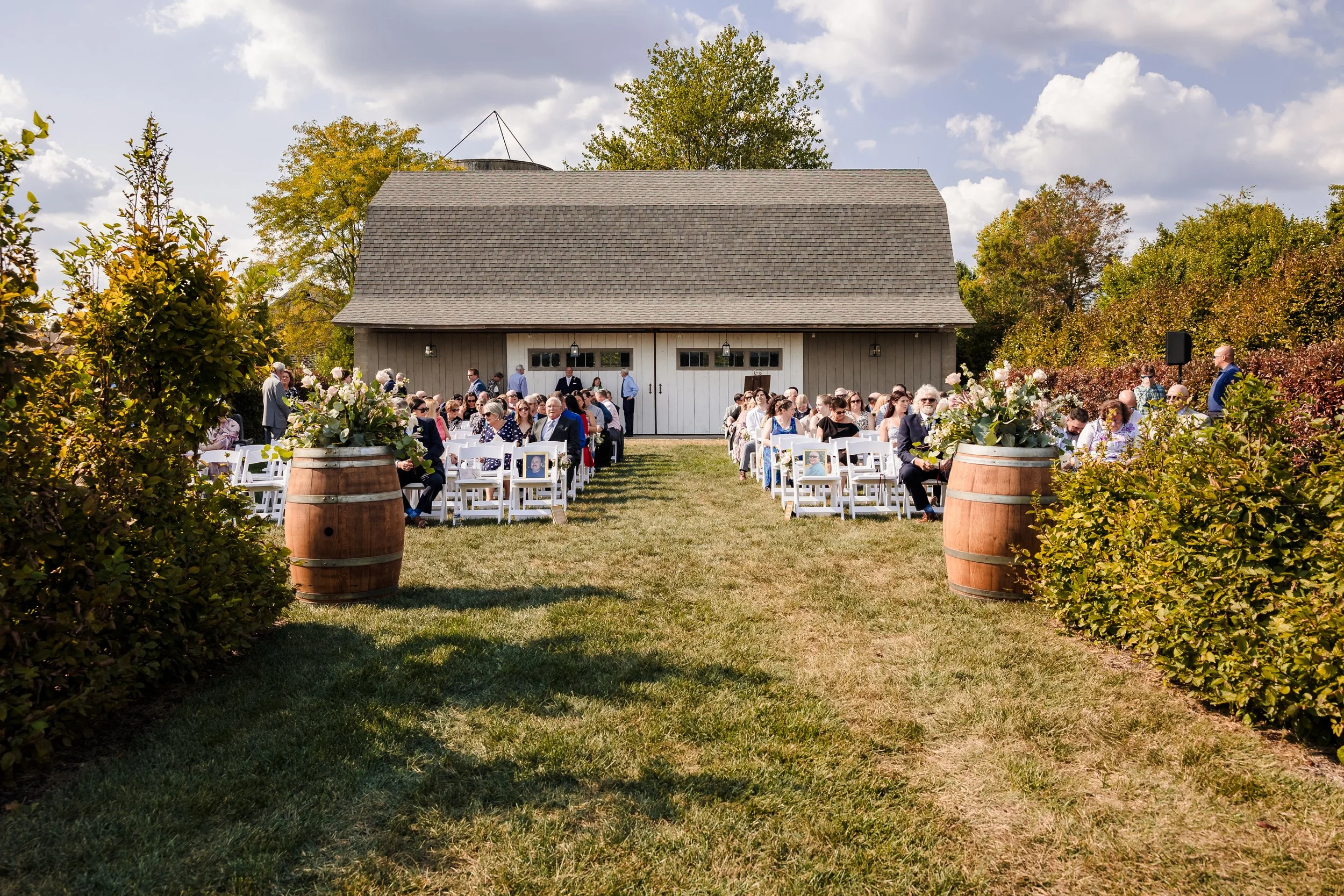 Guests seated on white chairs in front of a barn, outdoors during daytime, with trees and cloudy sky in the background, likely at a wedding or outdoor event.