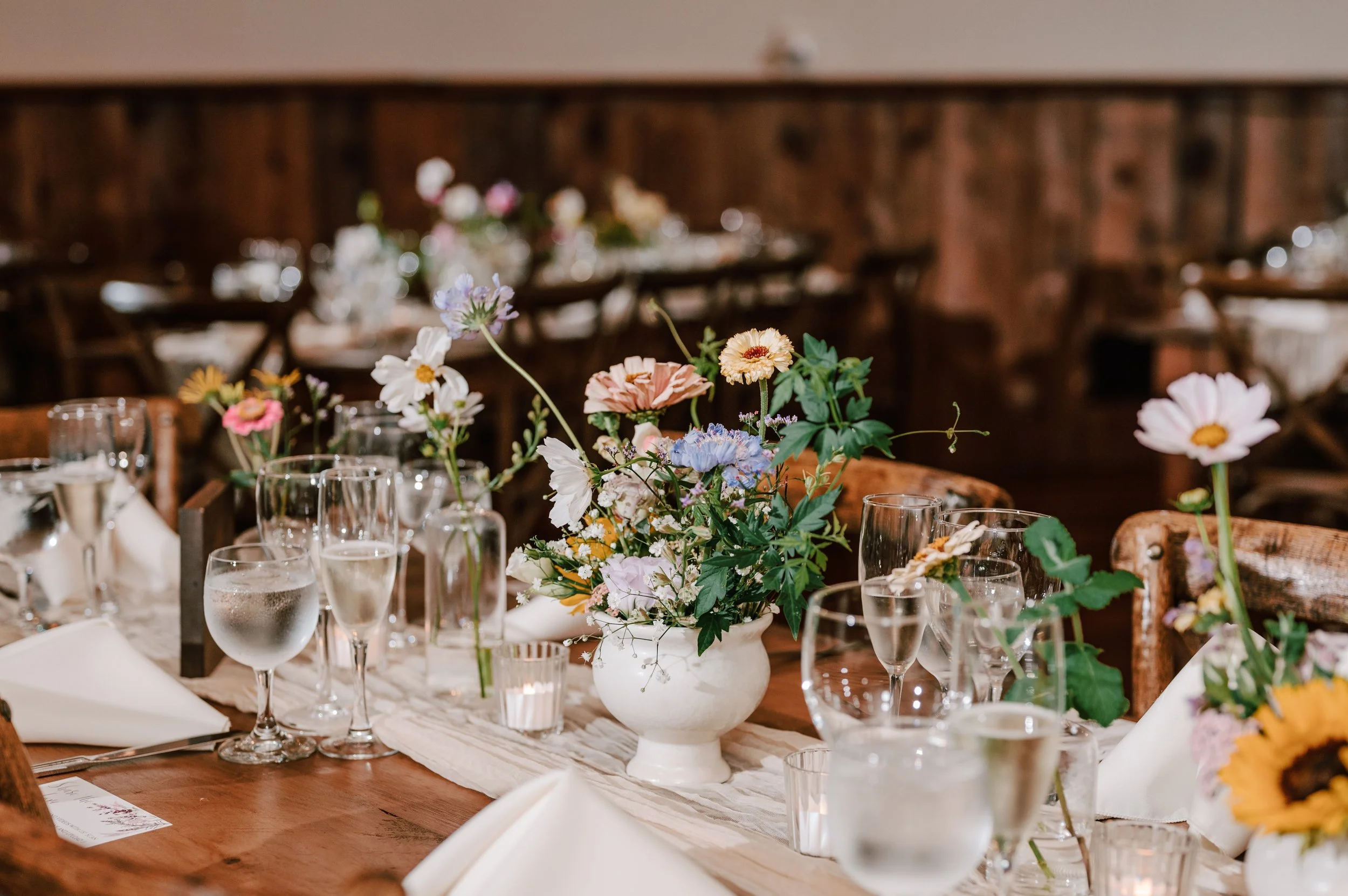Elegant dining table decorated with a variety of colorful flowers in white vases, several glasses, and white napkins, set for a special occasion inside a room with wood paneling.