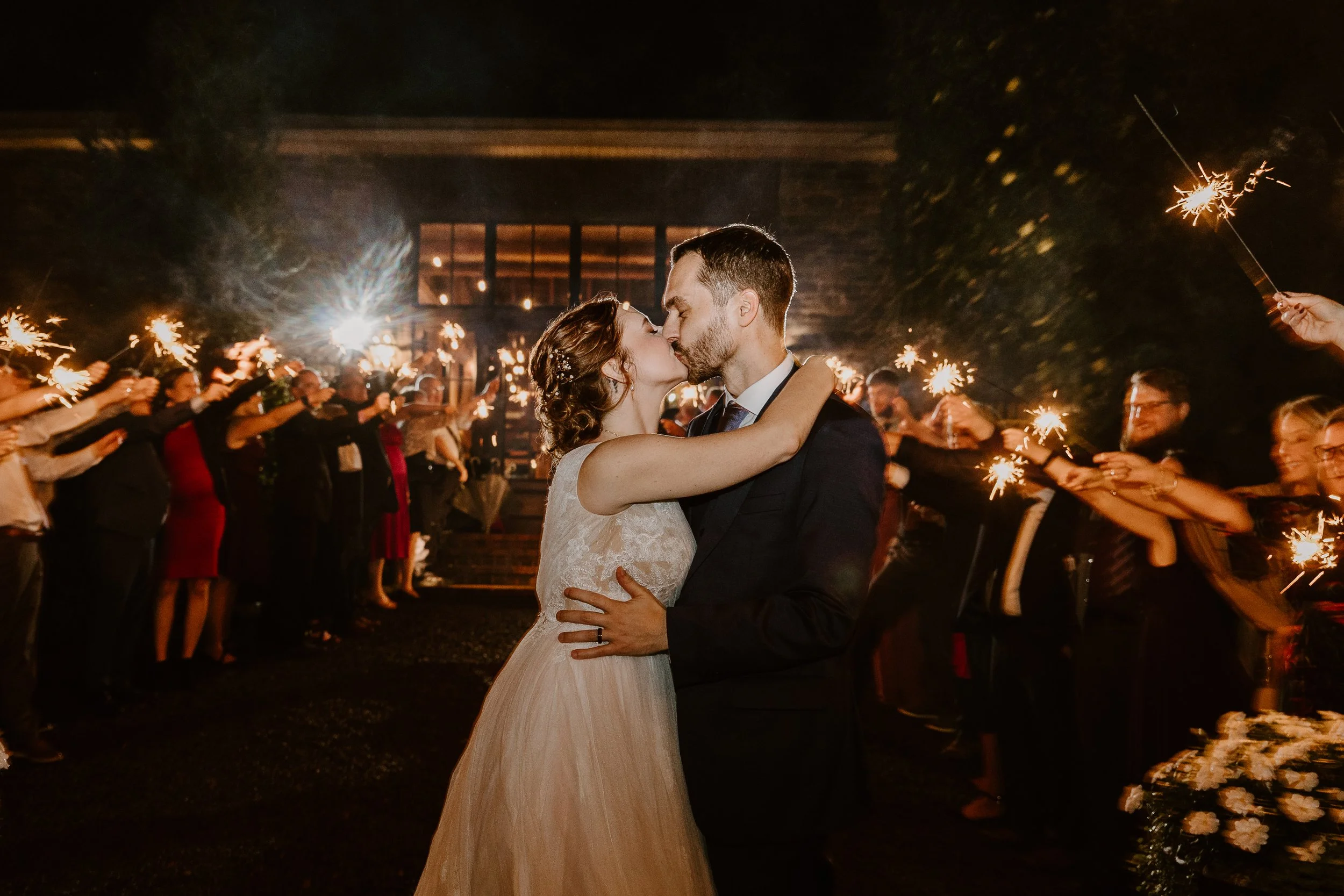A bride and groom kiss during their wedding celebration at night, with guests holding sparklers surrounding them, creating a festive and romantic atmosphere.