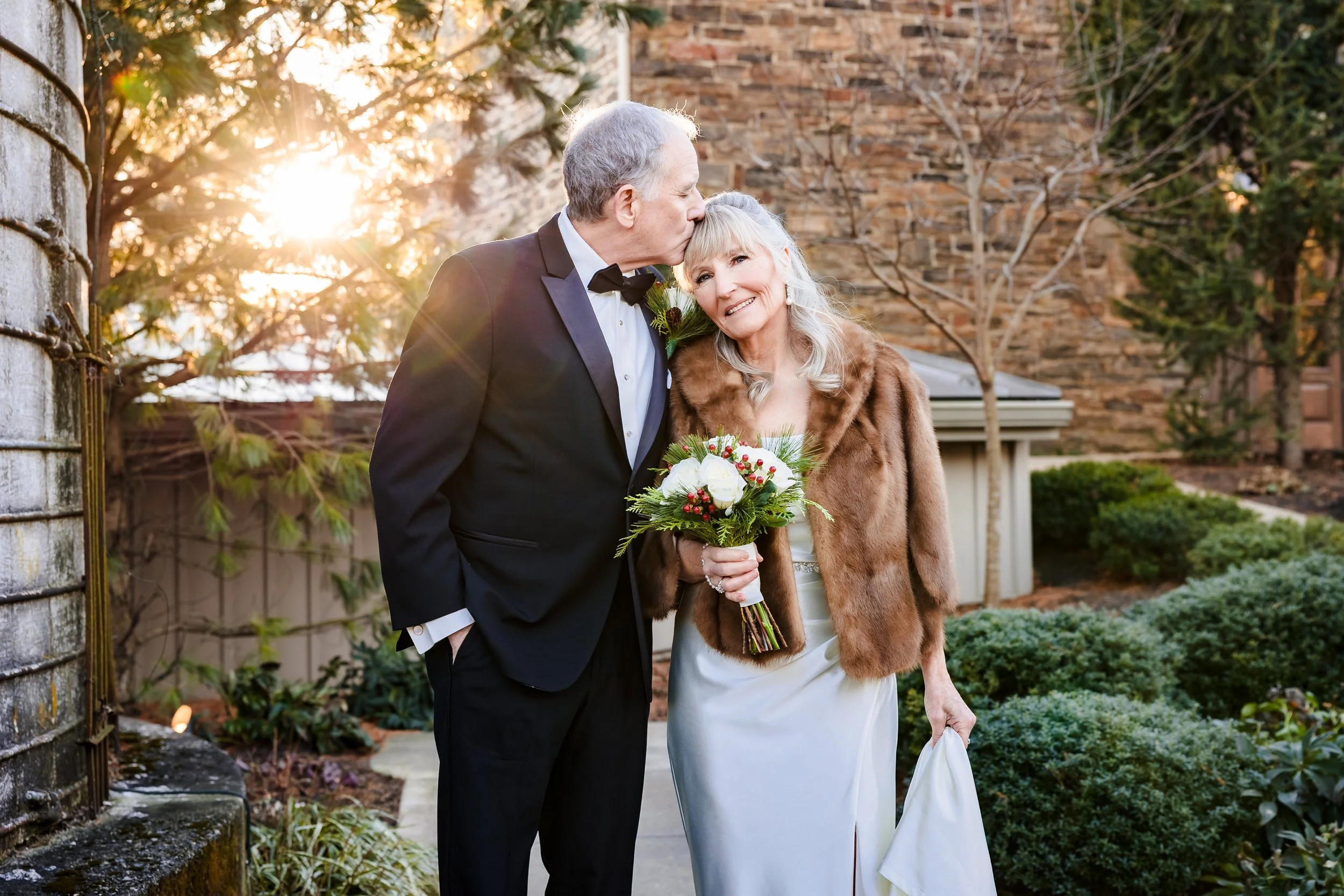 An elderly man in a tuxedo kisses an elderly woman in a white wedding dress and brown fur coat on the forehead during a wedding ceremony outdoors at sunset, with trees and a brick wall in the background.