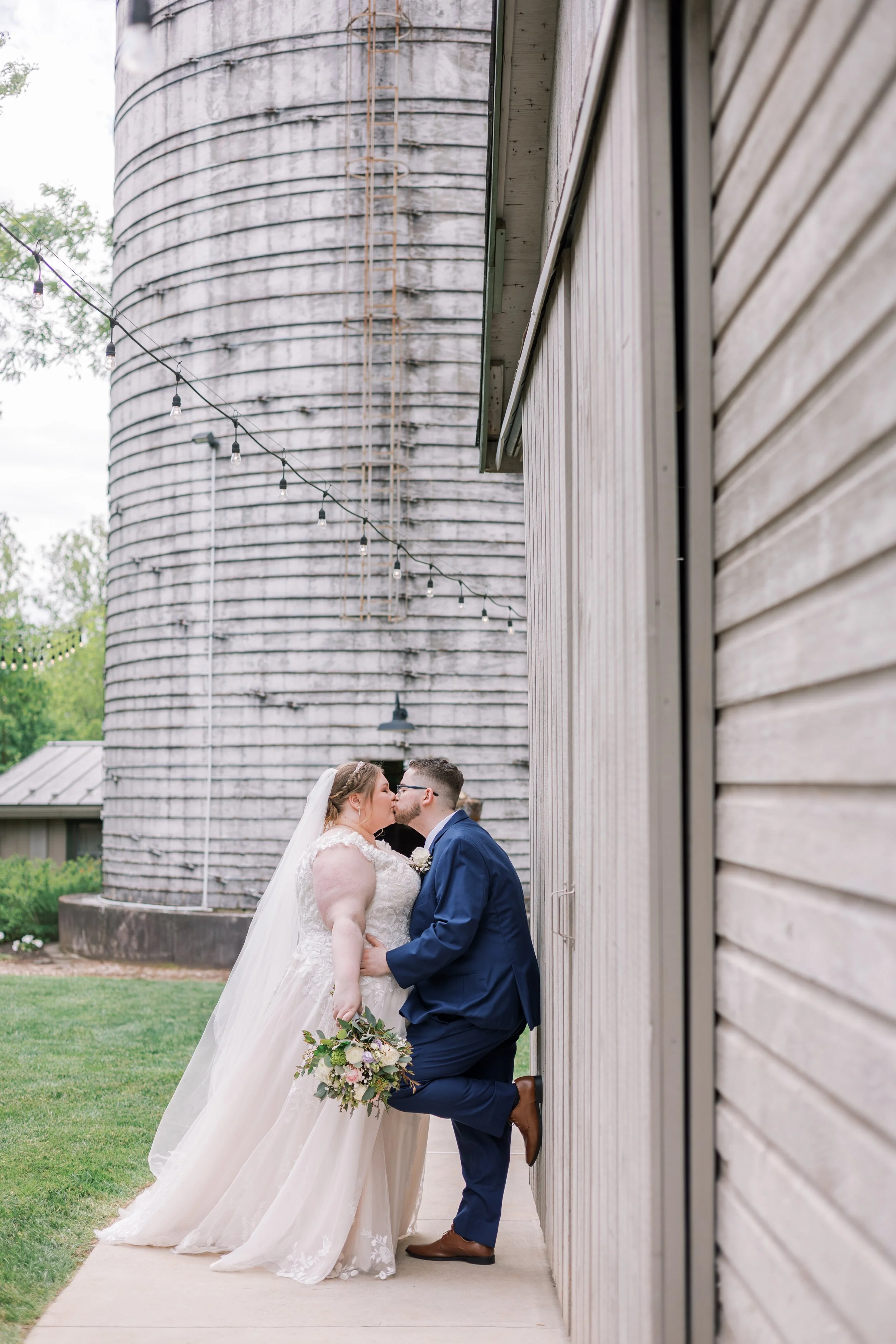 A bride and groom sharing a kiss outdoors during their wedding, with the bride holding a bouquet of flowers. They are standing close, with the groom leaning against a wooden wall, and a large wooden barn or silo visible in the background.