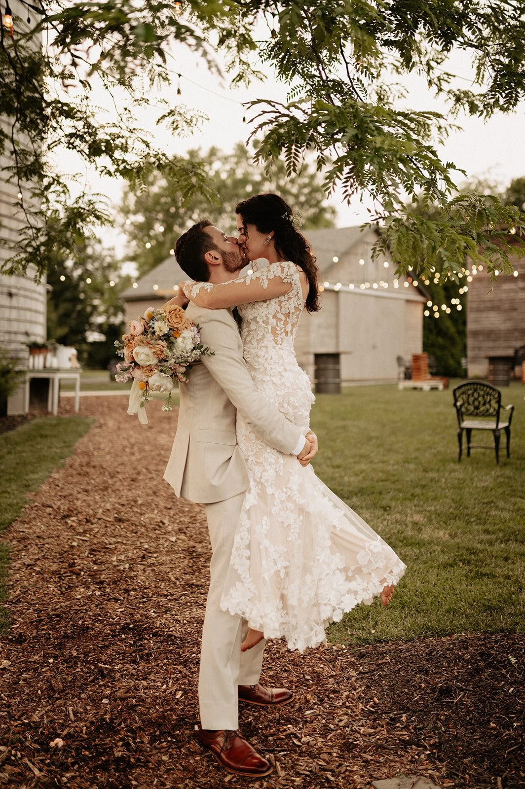A bride and groom share a kiss outdoors during sunset, with the groom lifting the bride while she holds a bouquet of flowers, surrounded by trees and rustic farm buildings.