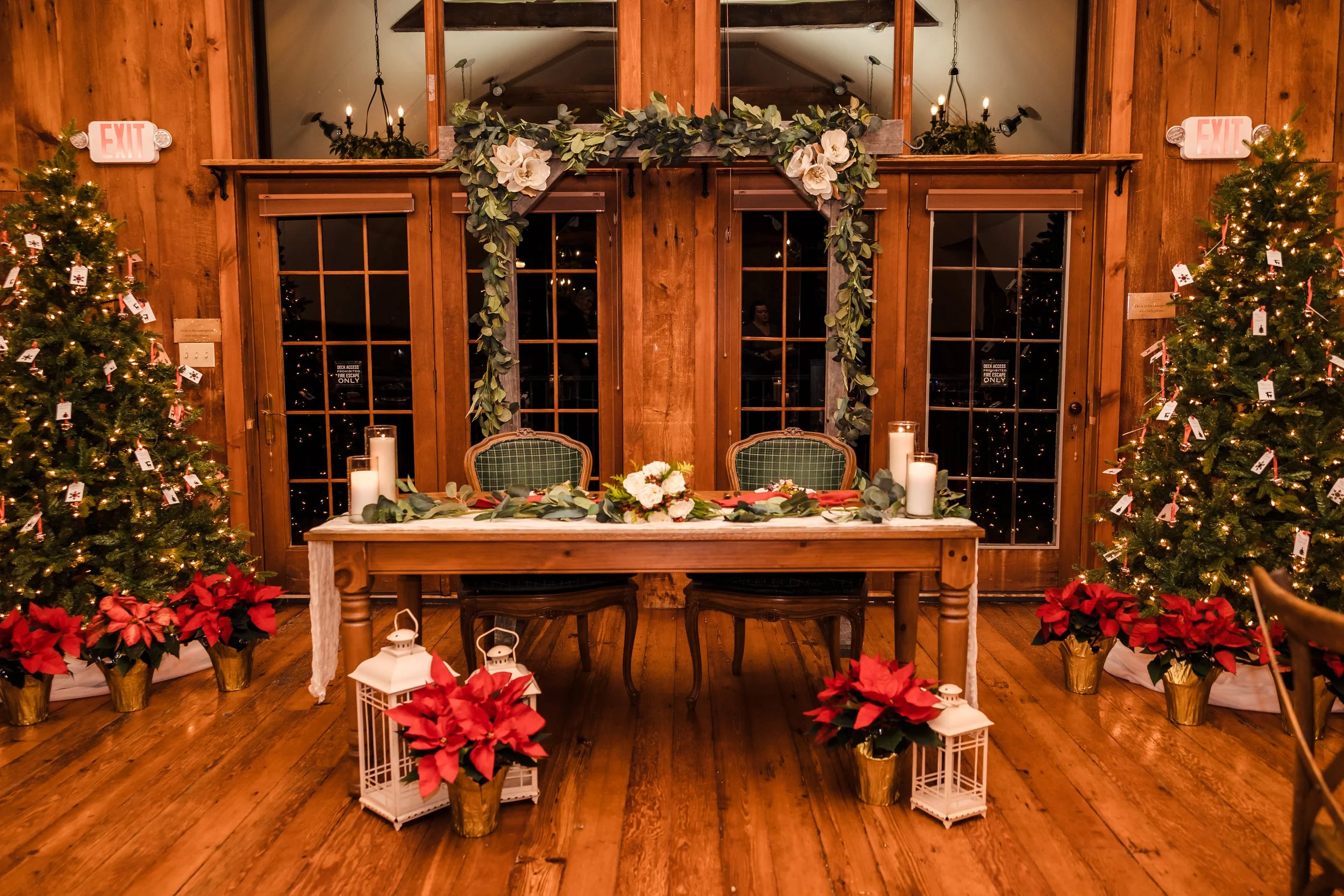 A festive Christmas scene featuring a wooden table with a poinsettia arrangement, candles, and lanterns in front of a wooden wall with decorated Christmas trees on either side, garland, and candles.