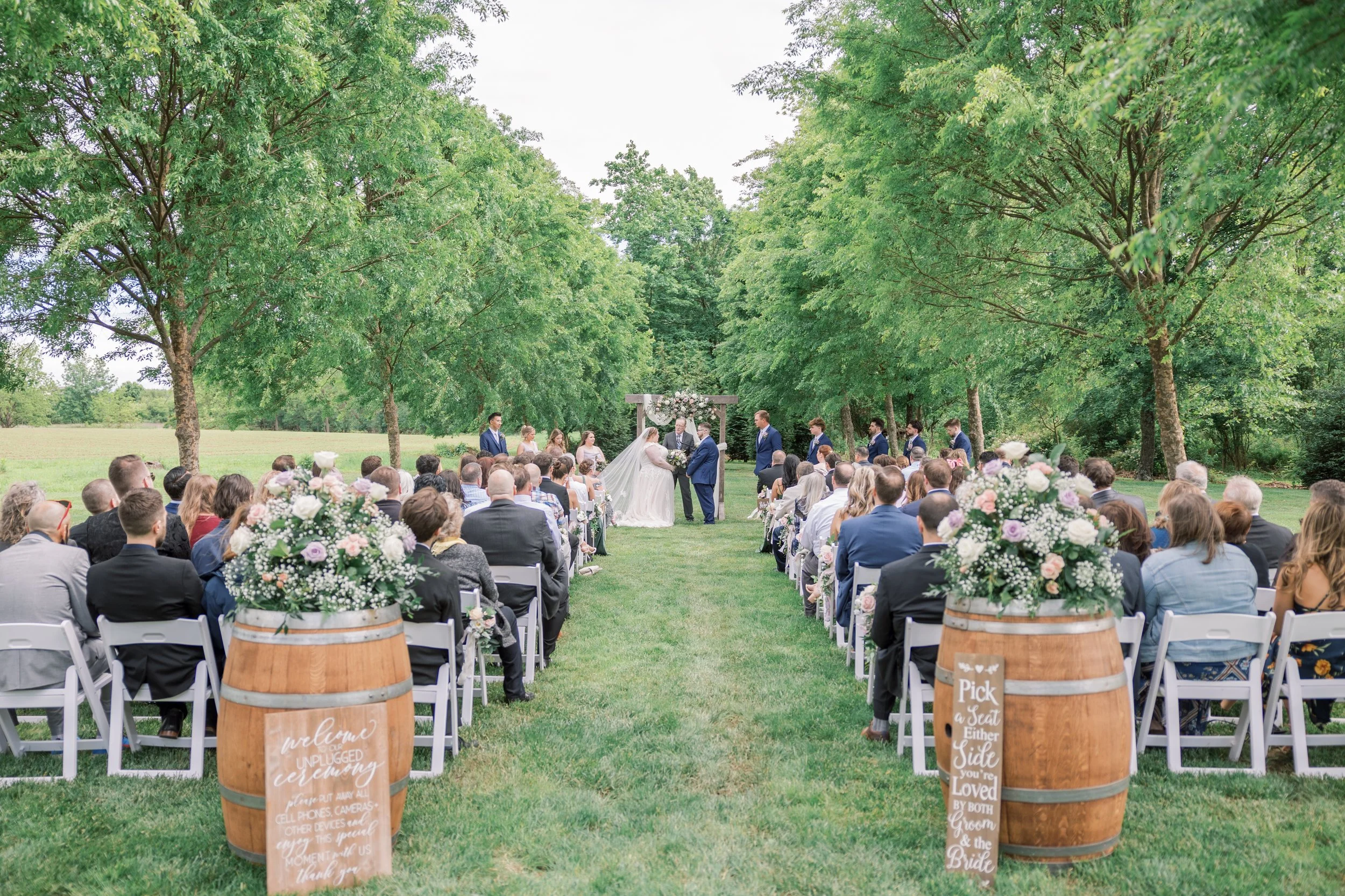 Outdoor wedding ceremony in a green park with guests seated on white chairs, floral arrangements, and a wooden arbor at the front.