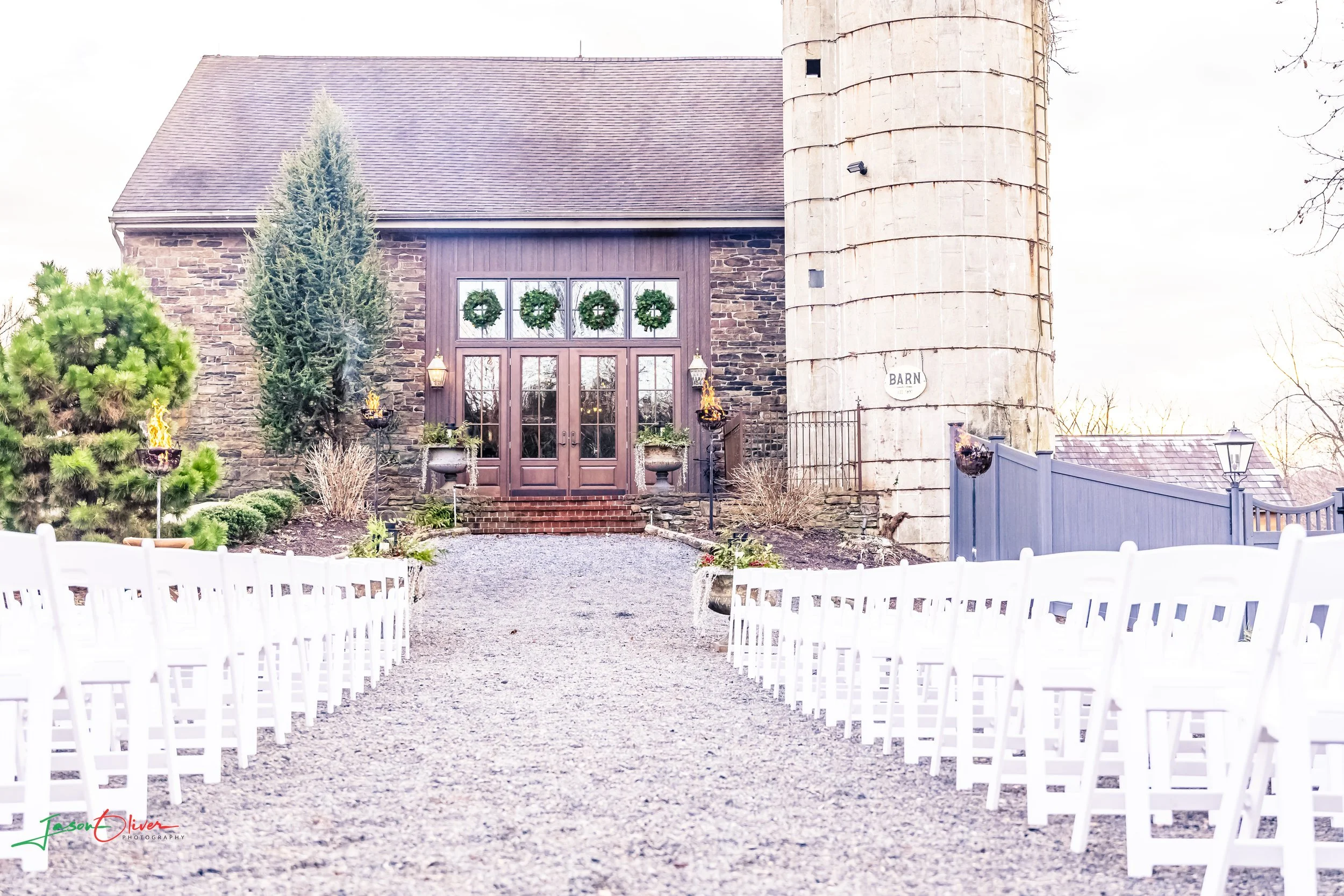 A rustic barn with a stone exterior and a brown wooden door decorated with four green wreaths. The barn is flanked by evergreen trees and a white picket fence. The entrance path is lined with small shrubs and outdoor lanterns, with a large silo on th