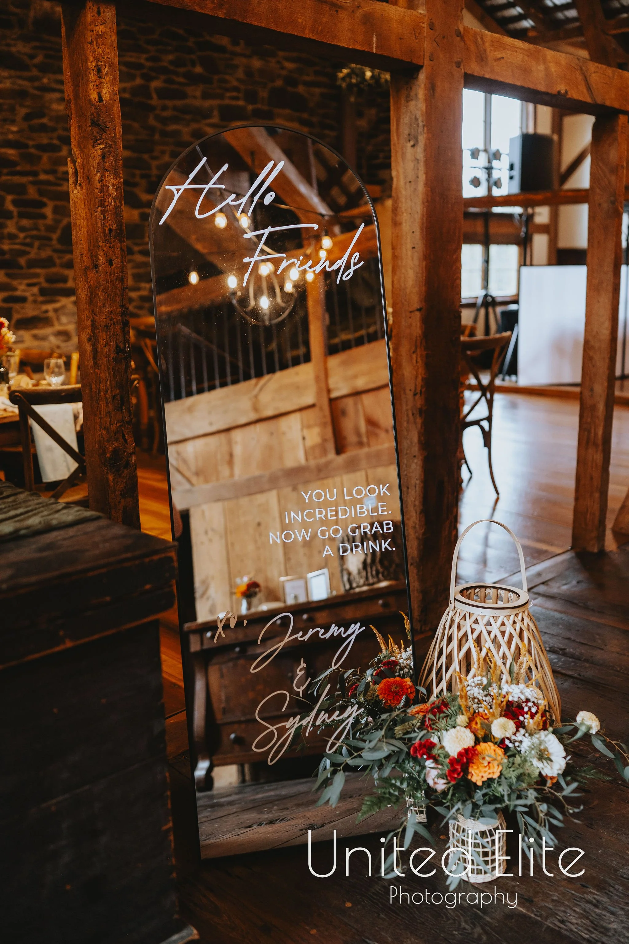 Decorative sign with the message 'Hello Friends' and additional text beneath, displayed in a rustic interior with wooden beams and a flower arrangement in a white lantern-style vase. The setting appears to be a cozy, rustic event or gathering venue.