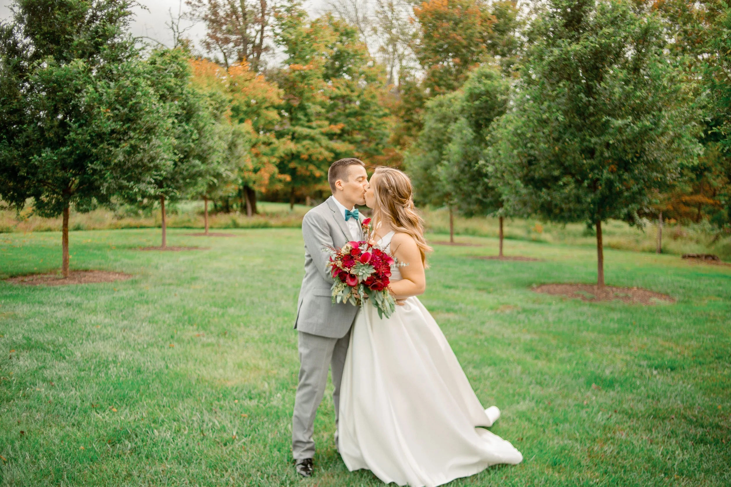 A bride and groom kissing outdoors on a grassy field with green trees in the background. The groom is wearing a light gray suit with a teal bow tie, and the bride is in a white wedding dress holding a red and pink bouquet.