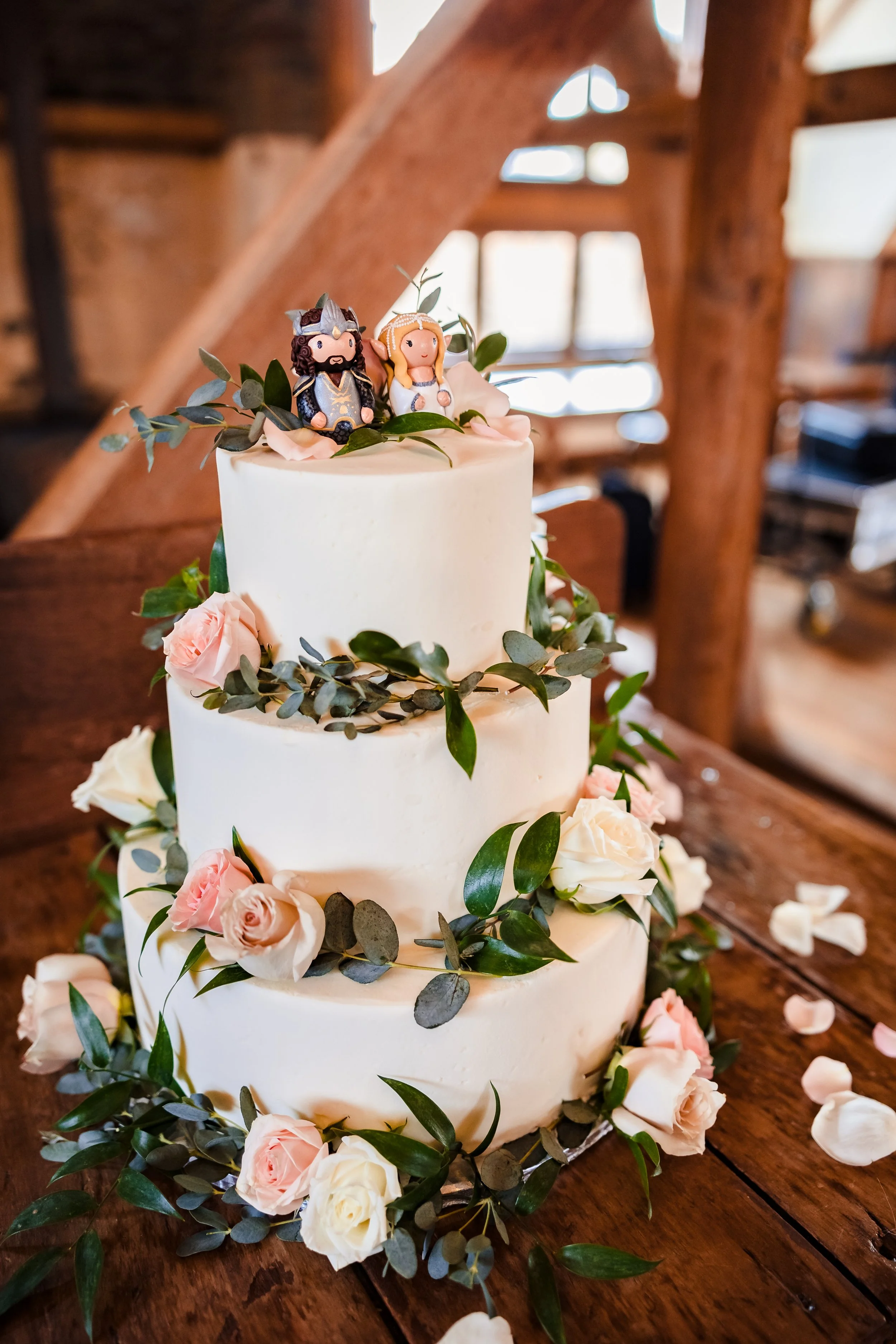 A four-tiered wedding cake decorated with pink and white roses, green leaves, and small figurines of a bride and groom on top, placed on a wooden table.