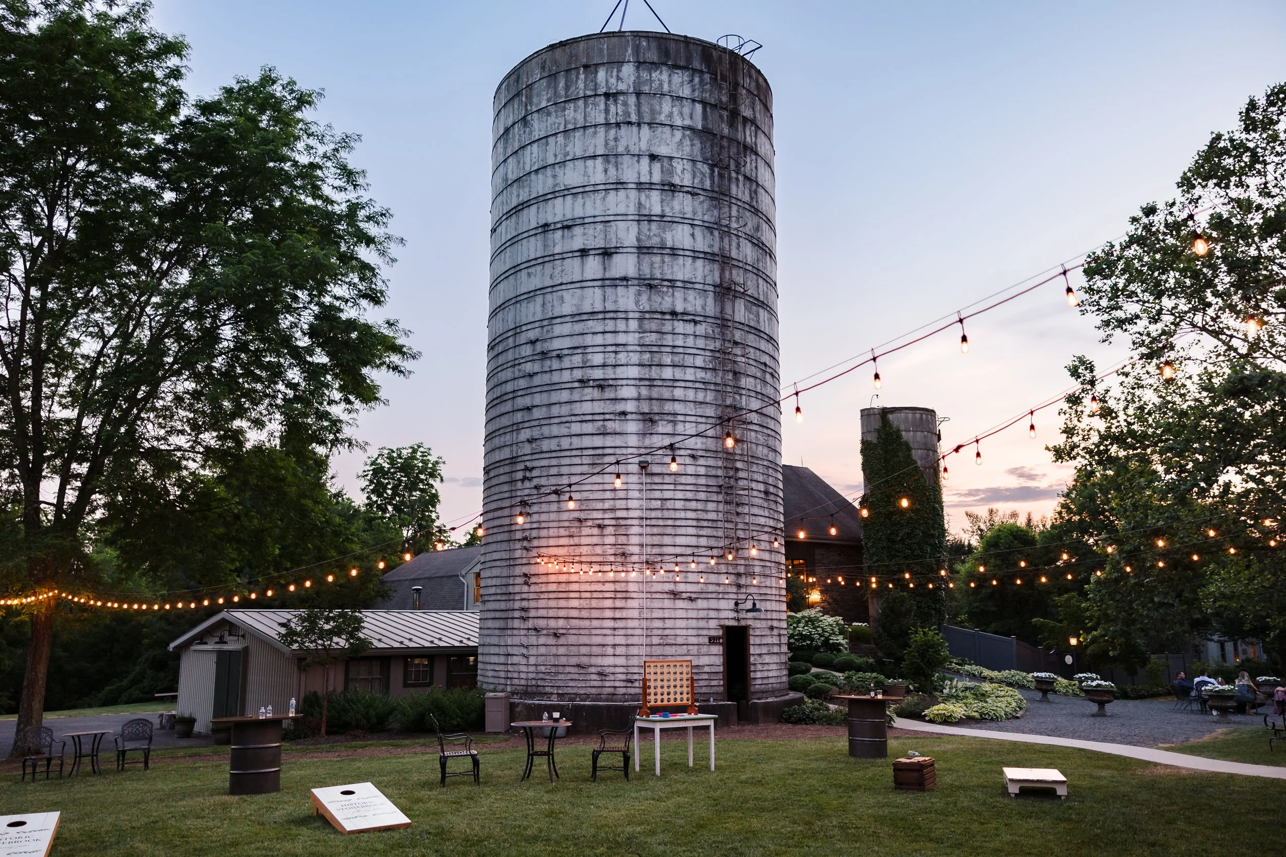 A tall, cylindrical, weathered barn silo surrounded by large trees, string lights, and outdoor tables and chairs in a garden setting during early evening.
