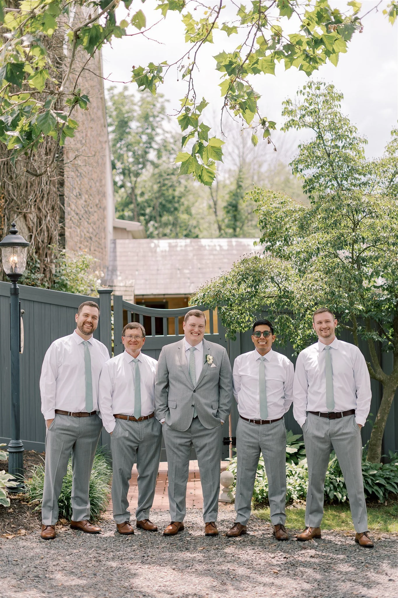 A group of five men dressed in suits, posed outdoors in front of a garden fence, with trees and a brick building in the background.