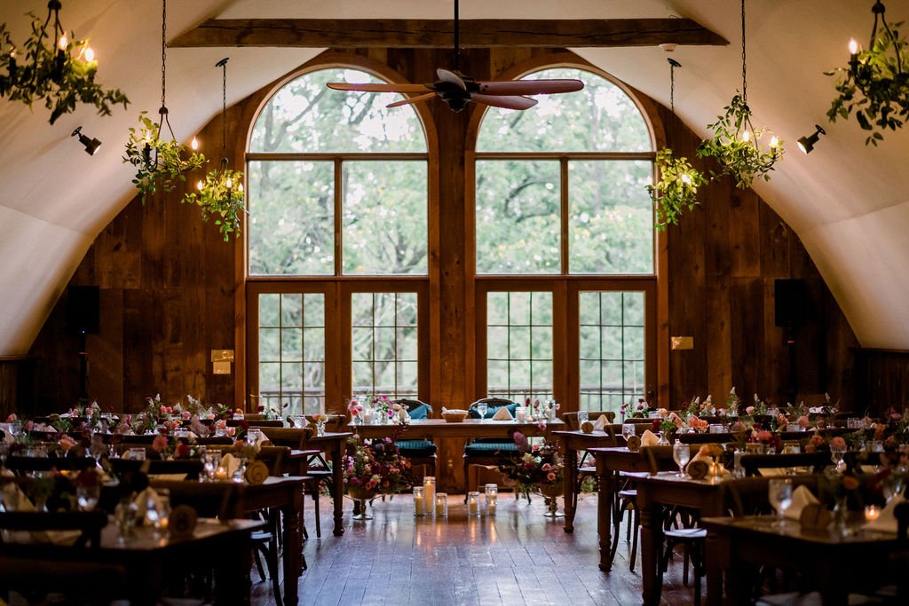 Elegant event space with wooden walls and large arched windows, decorated with hanging plants, flowers, candles, and neatly arranged tables, illuminated by warm lighting.