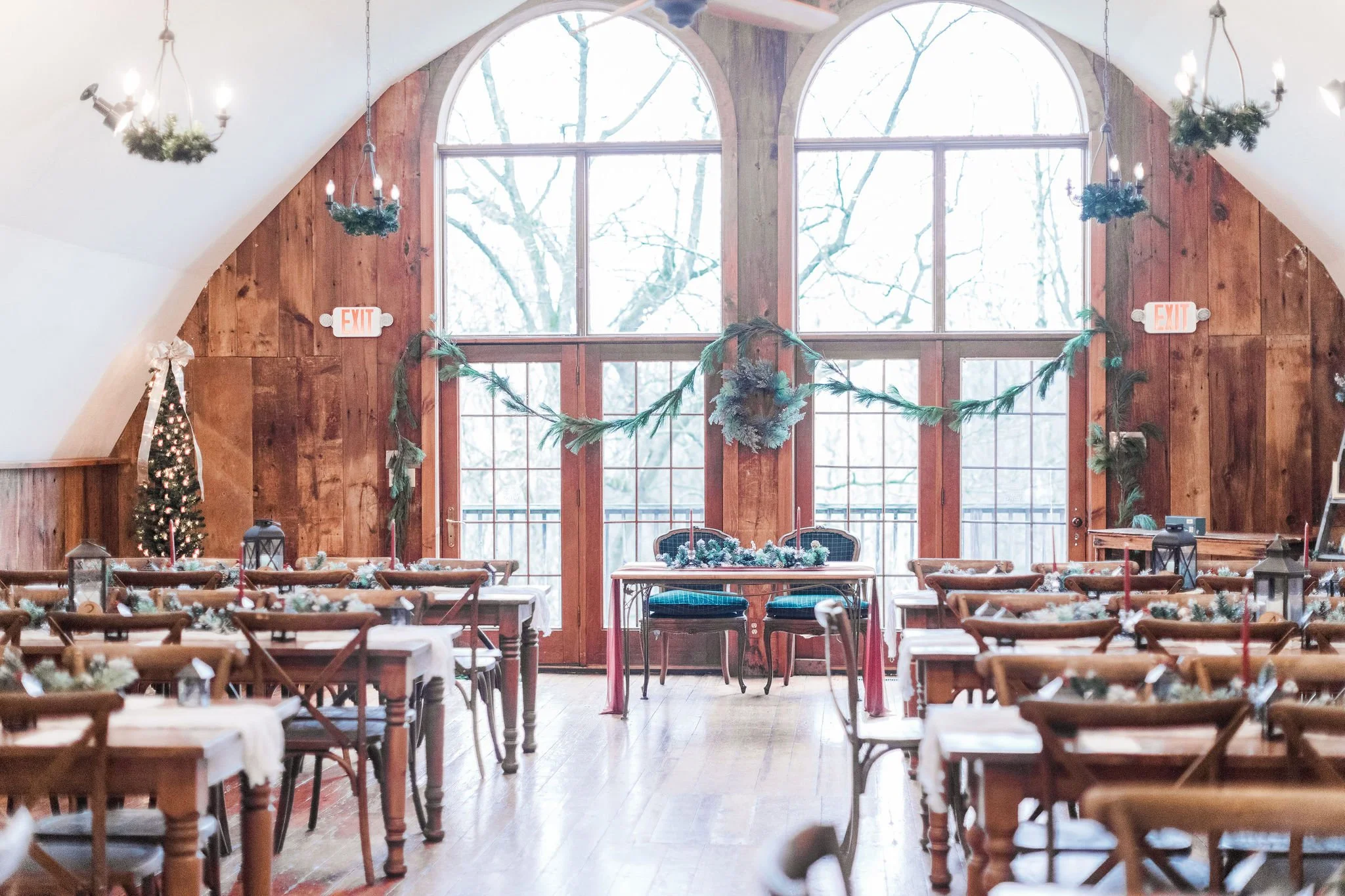 A decorated dining hall with wooden walls and large arched windows, featuring Christmas garlands, candles, and a small Christmas tree with lights.