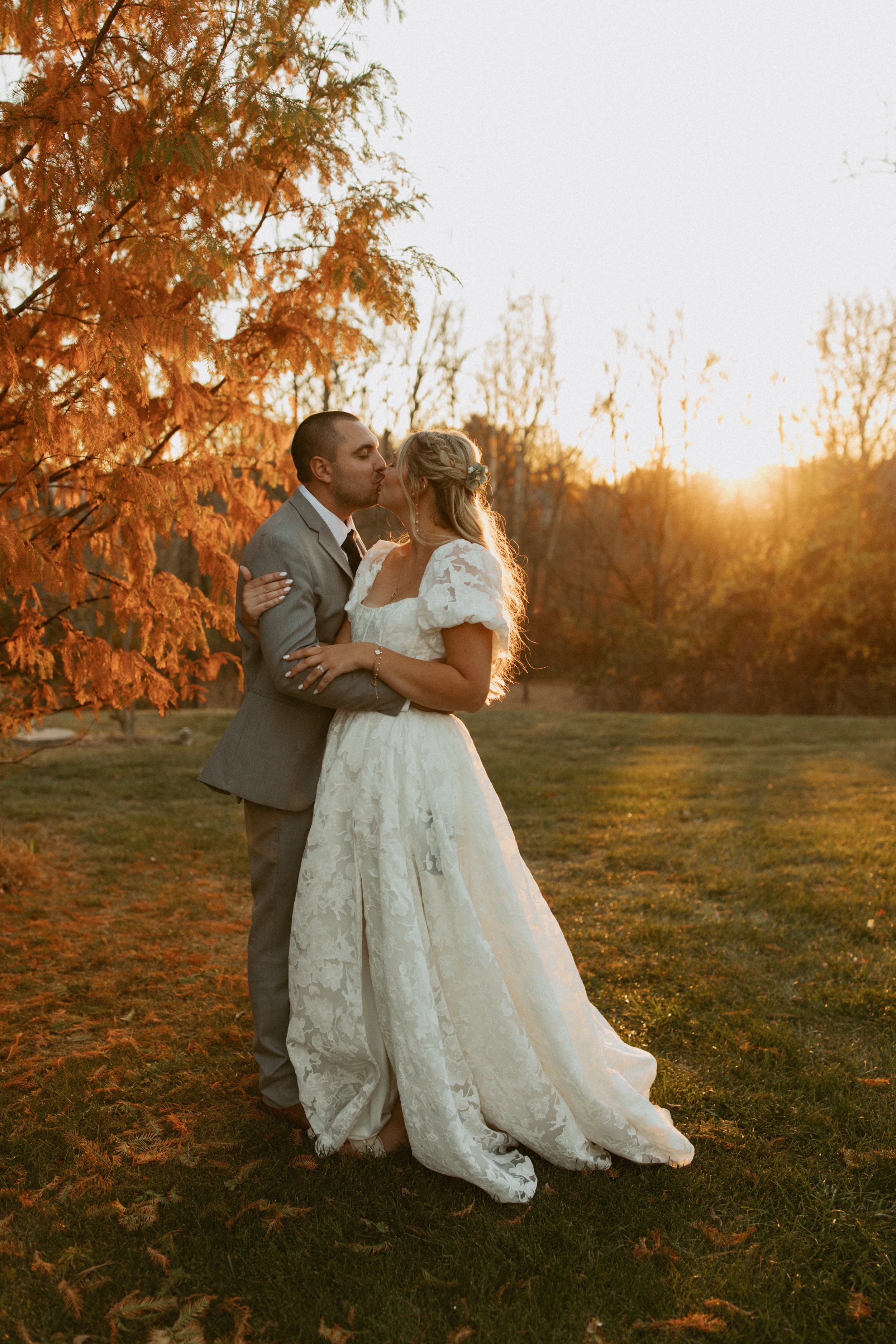 A newlywed couple sharing a kiss outdoors during sunset in an autumn setting with orange leaves and trees in the background.