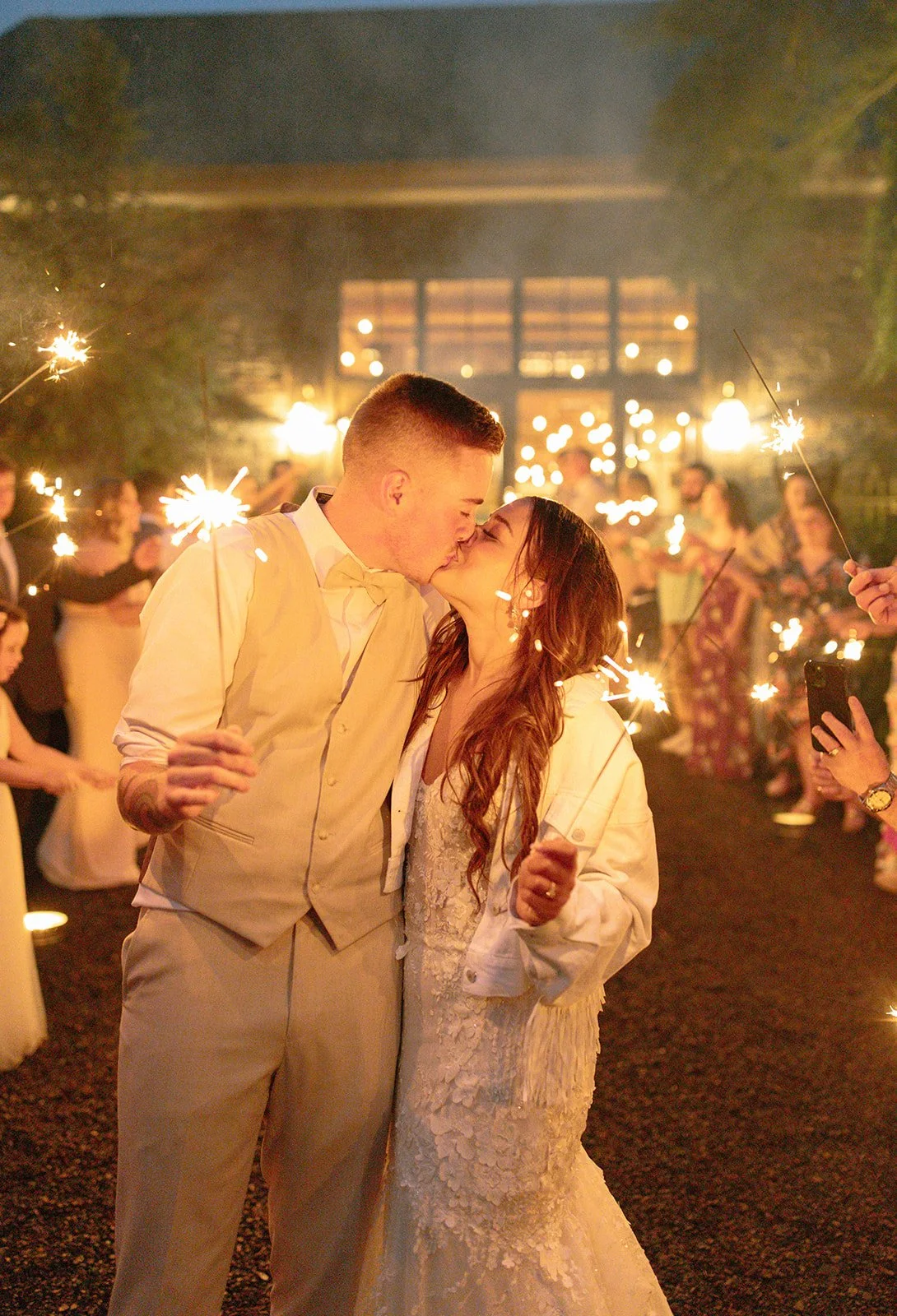 A couple kissing at a nighttime celebration with sparklers, surrounded by guests holding sparklers at an outdoor event.