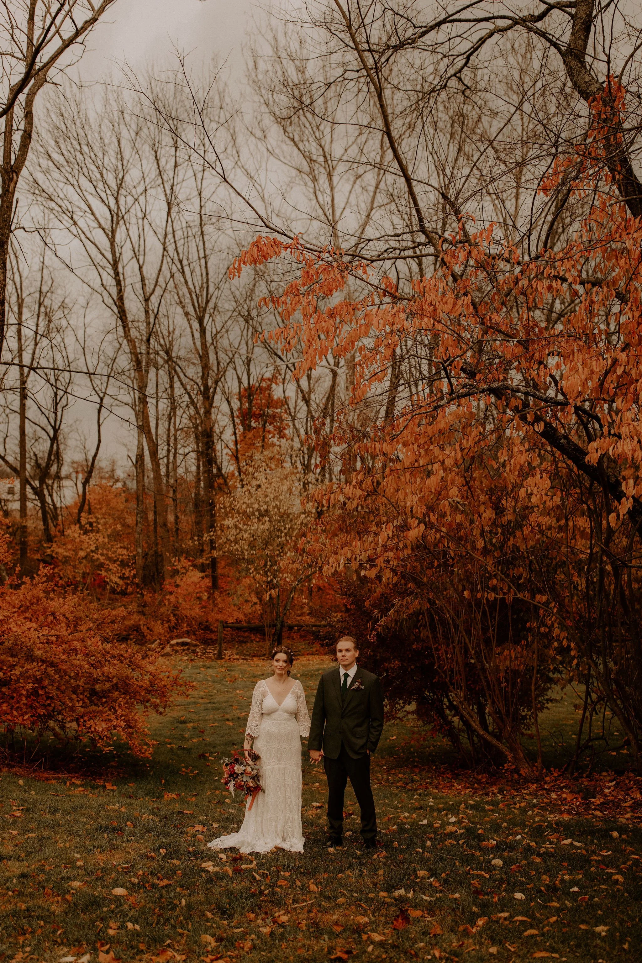 A bride and groom holding hands, walking outside in a park during fall, with orange and brown leaves and trees with bare branches in the background.