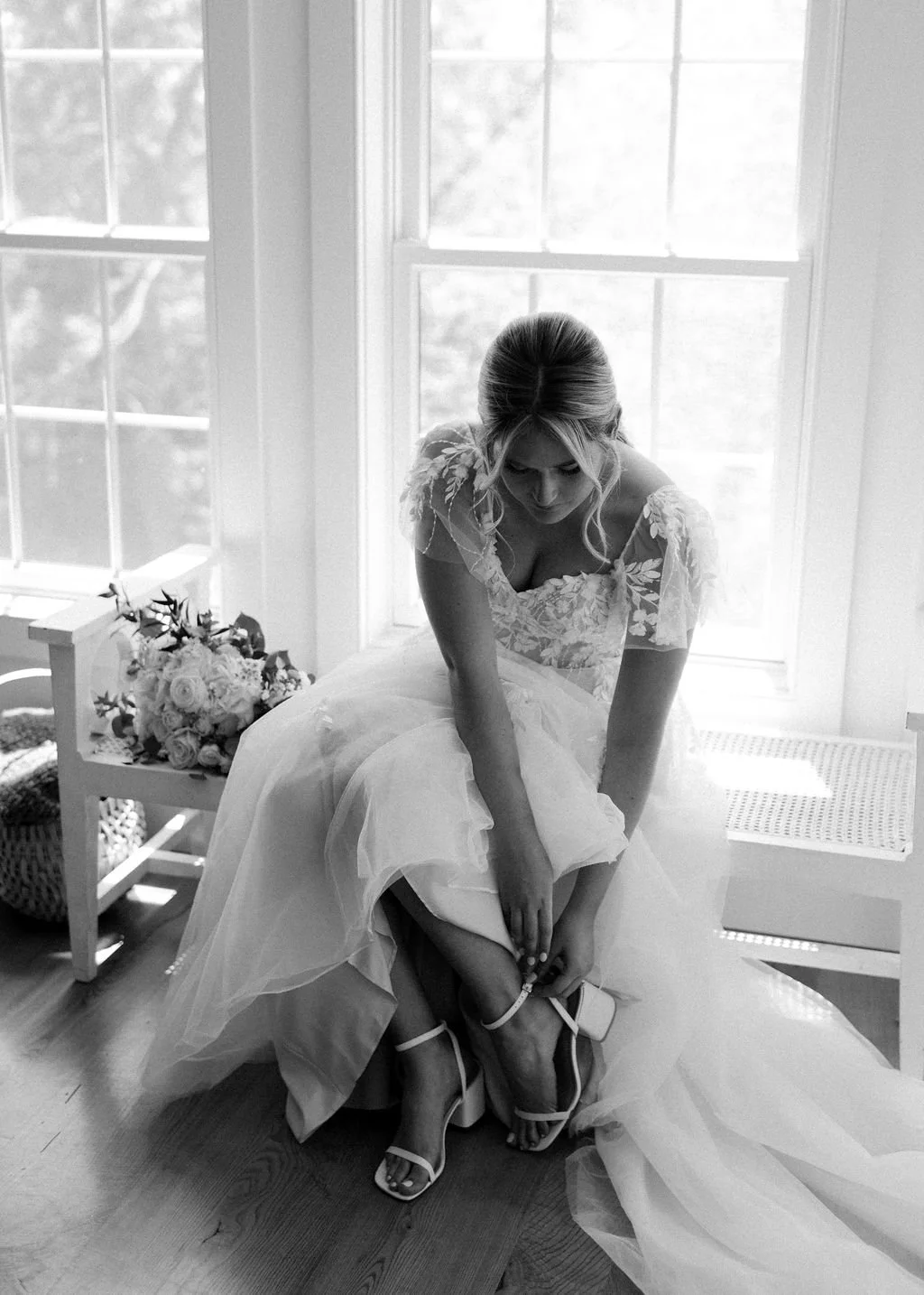 A bride sitting on a bench near a window, adjusting her high-heeled shoes, with a bouquet of flowers nearby, in a bright room.