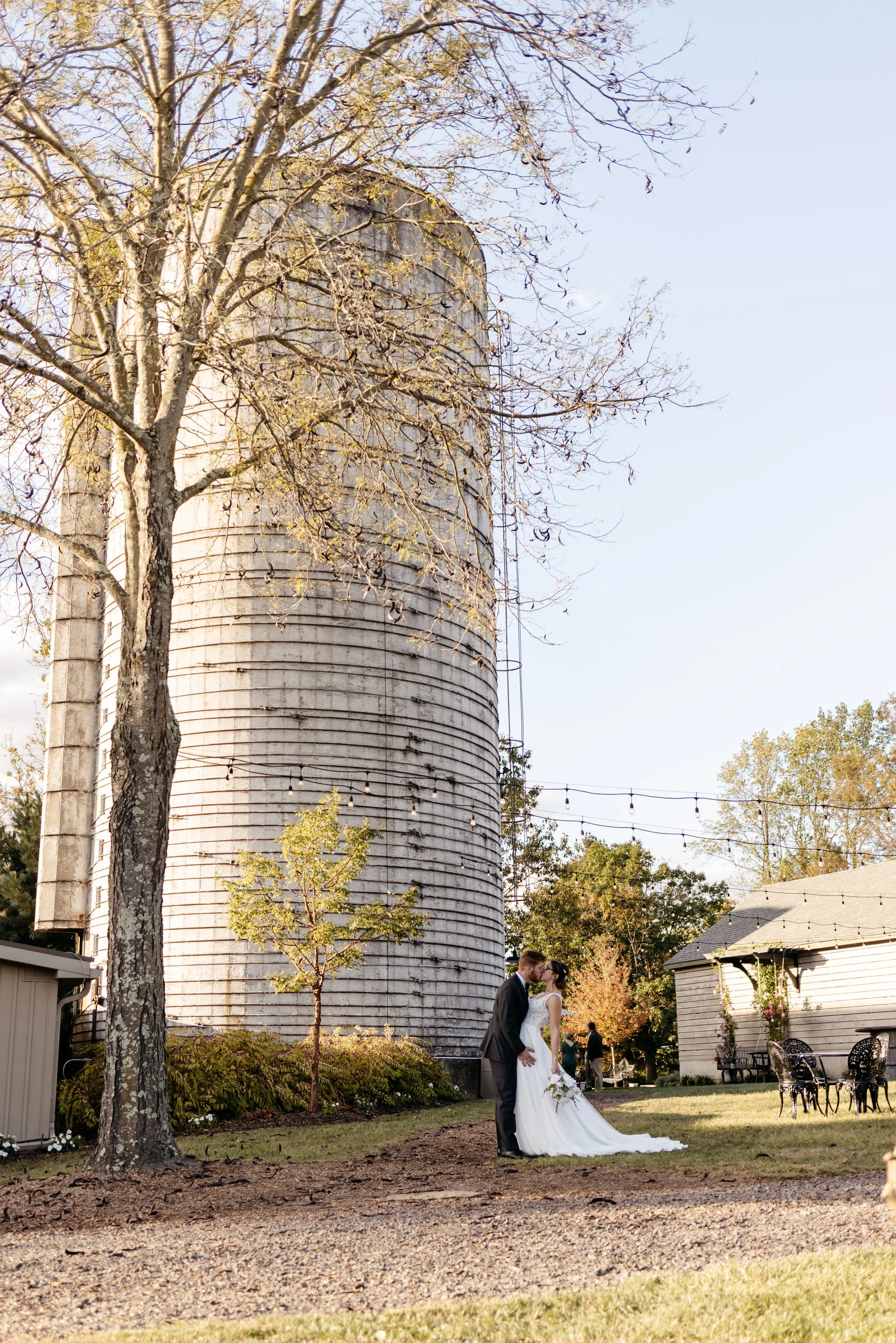A bride and groom kiss outdoors in a garden area with string lights, trees, a large silos, and a small building in the background, during what appears to be a wedding celebration.
