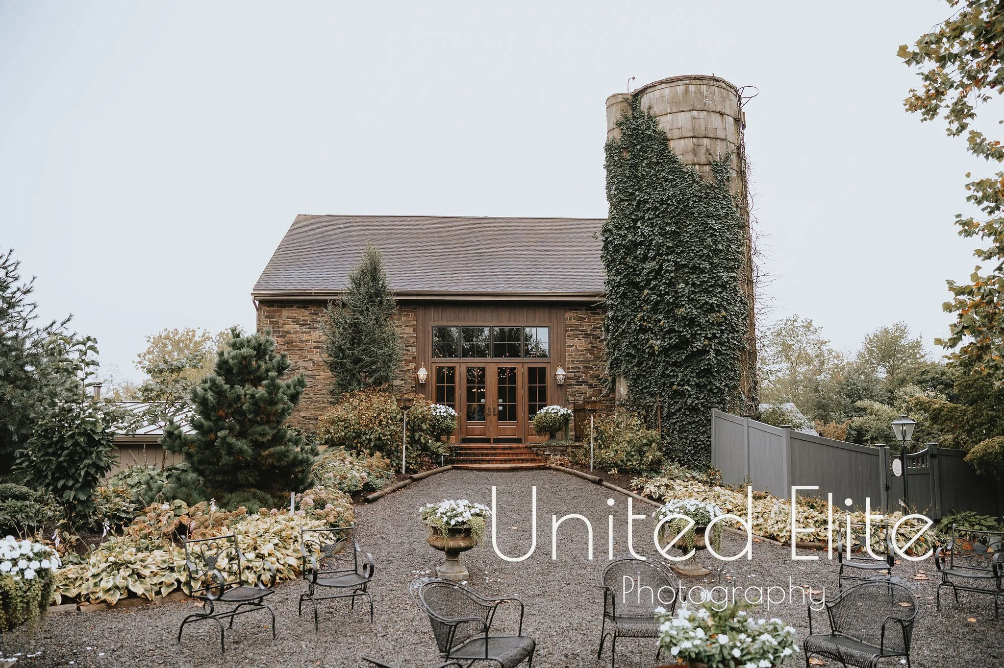 A rustic stone house with a wooden front door and large windows, surrounded by a garden with white flowers, small trees, and black metal chairs, with a tall, ivy-covered chimney rising above the house and cloudy sky in the background.