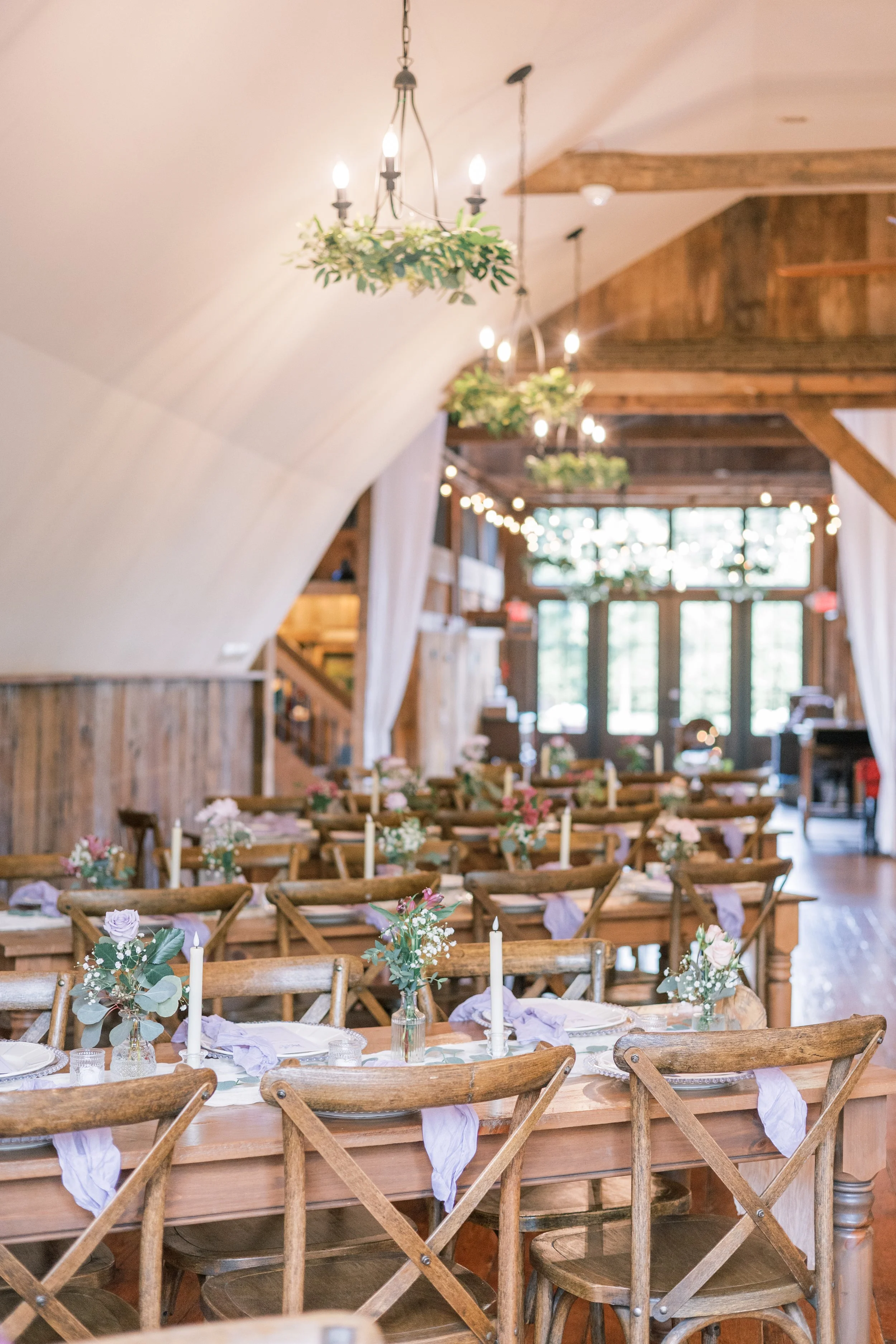 Indoor wedding reception with long wooden tables decorated with flower arrangements and candles, rustic wooden chairs, hanging chandeliers, and string lights in a barn-style venue with wood beams and large windows.