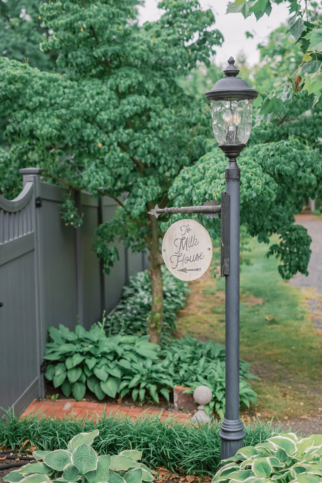 A decorative outdoor lamp post with a sign hanging underneath reading 'To Milk House' in a garden with lush green plants, a fence, and trees.