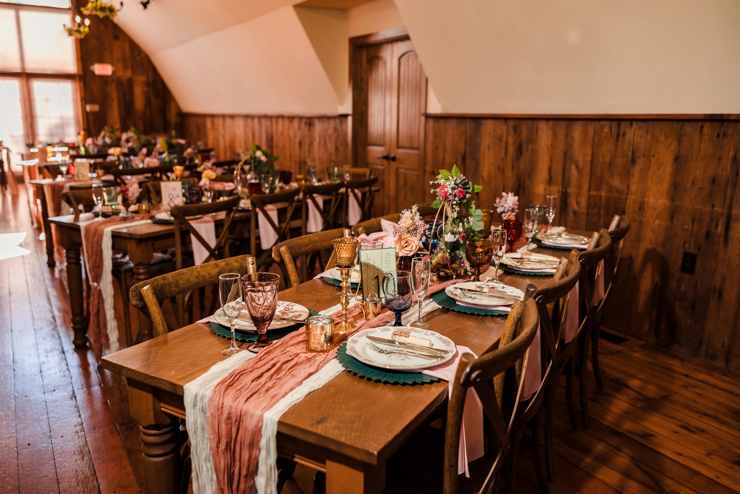 A decorated dining table set for a celebration in a rustic wooden room, with floral centerpieces, plates, glasses, and a pink and white table runner.