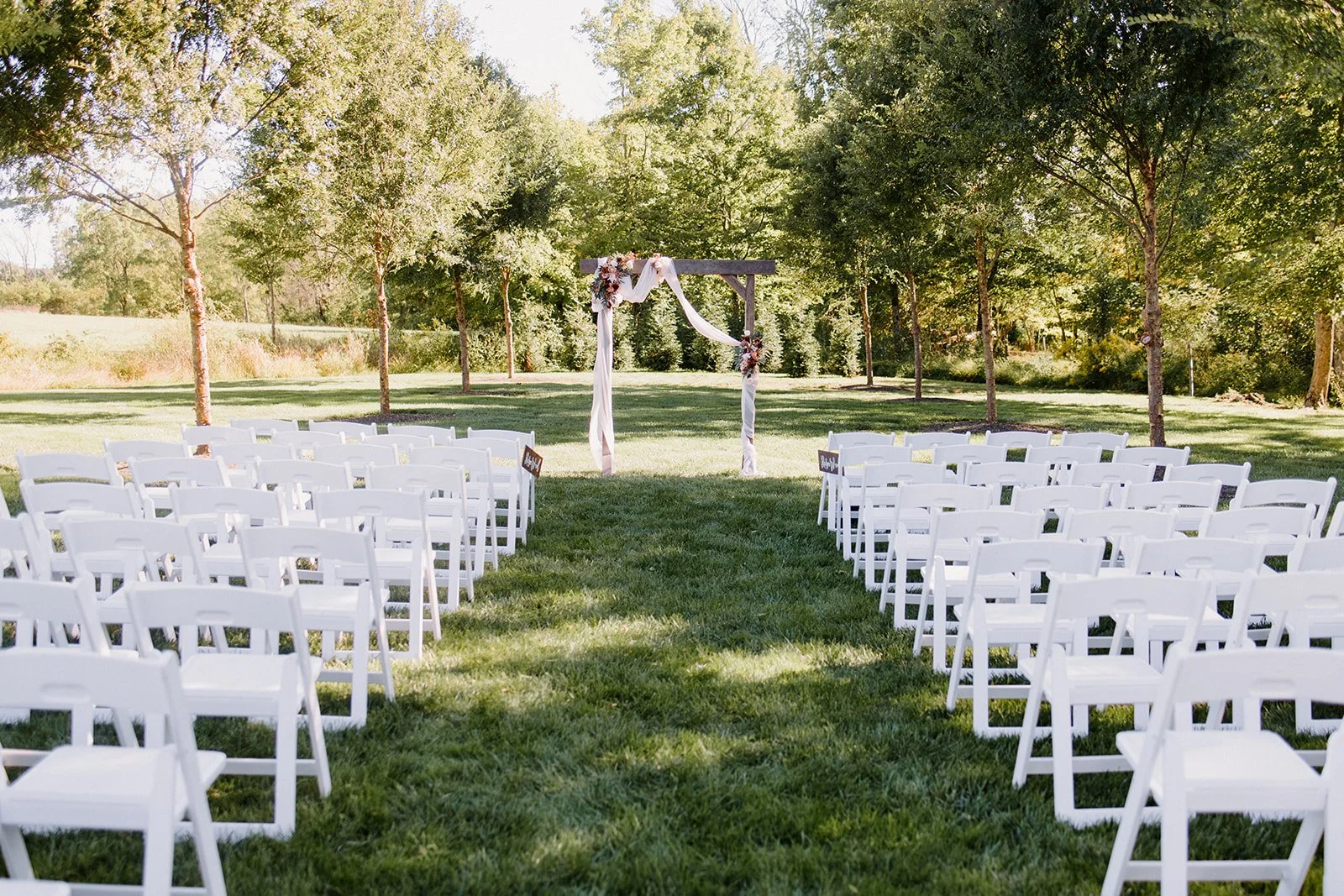 Outdoor wedding ceremony setup with white chairs arranged on green grass facing a wooden arch decorated with flowers and fabric, surrounded by trees on a sunny day.