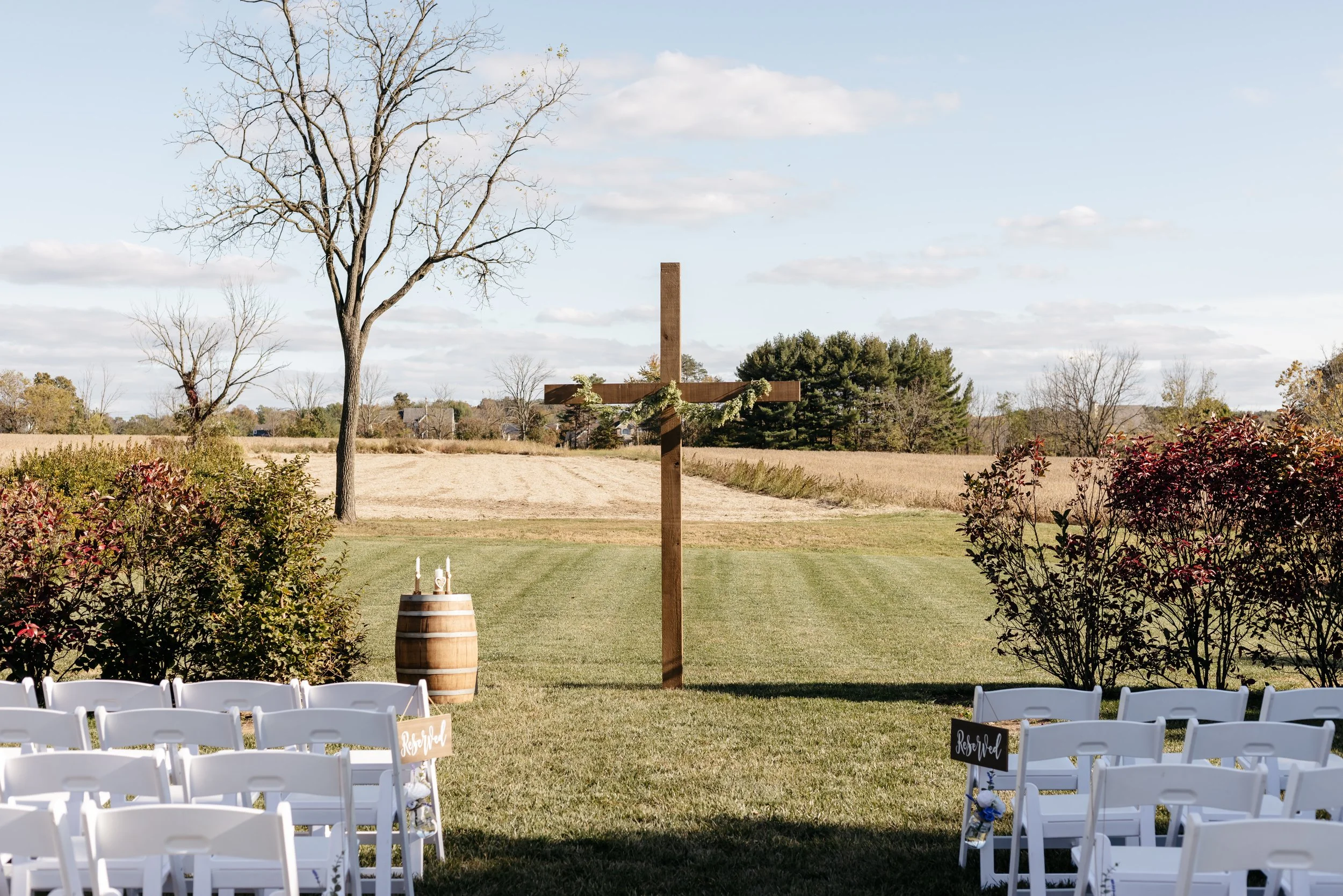 Outdoor wedding ceremony setup with white chairs, a wooden cross with greenery, a barrel with candles, and chairs marked 'Reserved' in a field with trees and a blue sky.